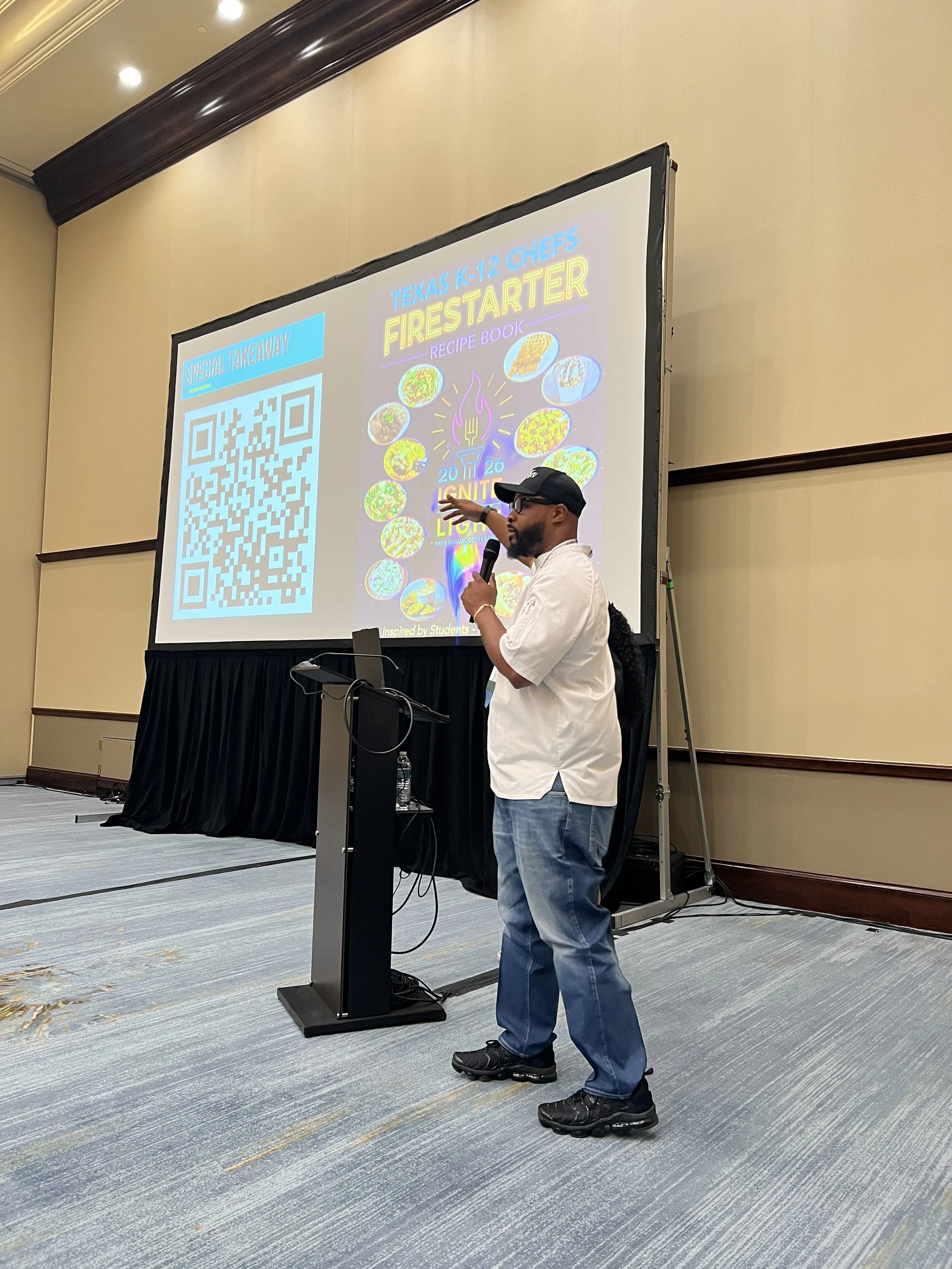A man with a beard, glasses, black cap, white shirt, and blue jeans stands at a podium speaking into a microphone. Behind him is a large projector screen displaying a colorful presentation slide with the words 'Texas K-12 Chefs Firestarter Recipe Boo