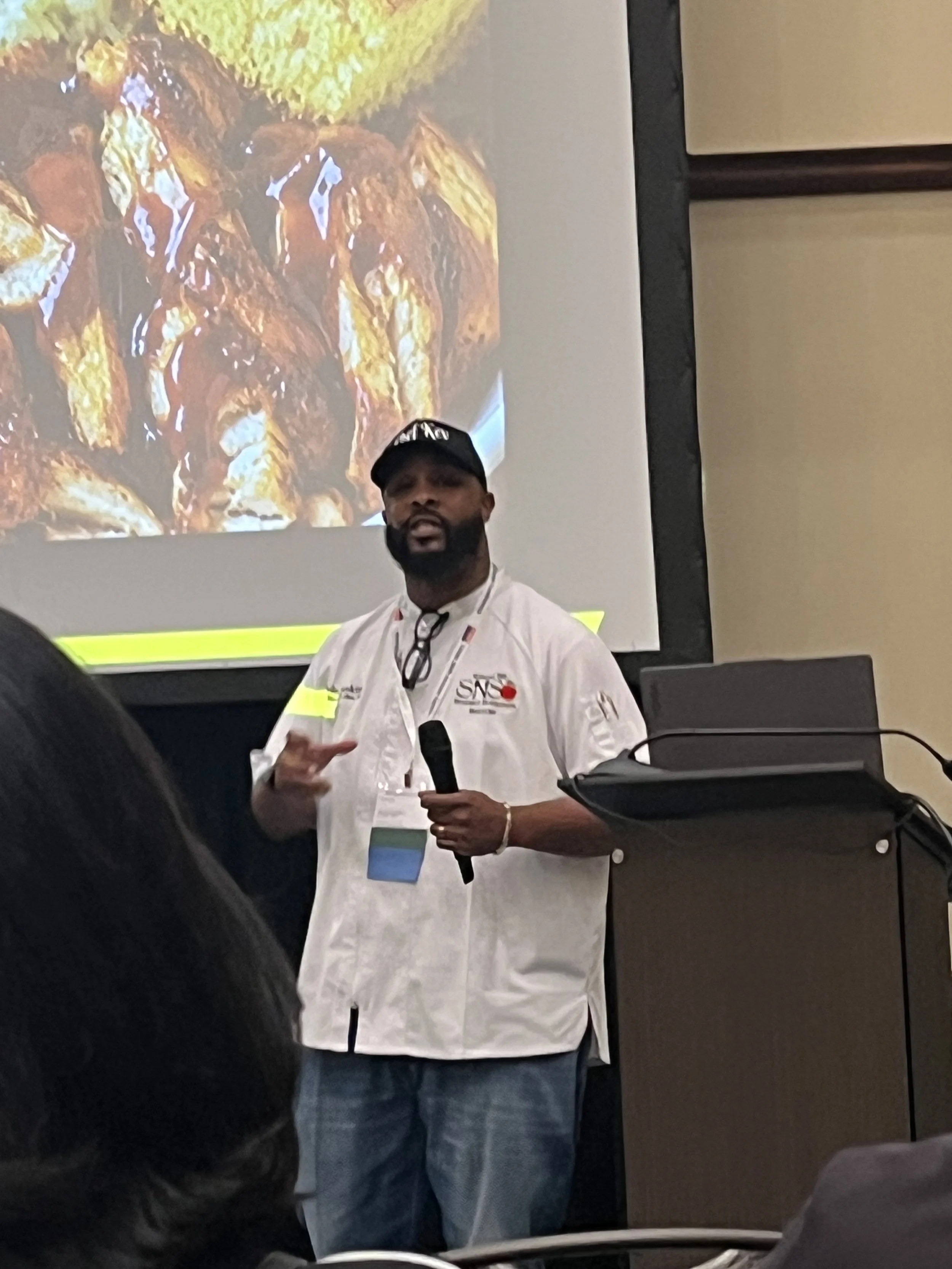 A man with a beard and wearing a black cap and white chef's jacket is speaking into a microphone. He is in front of a large screen displaying an image of fried chicken and lime wedges.