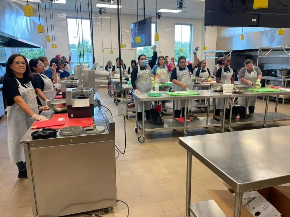 Group of people in a commercial kitchen participating in a cooking class, wearing aprons and gloves, some raising their hands, with kitchen appliances and utensils on the counters.