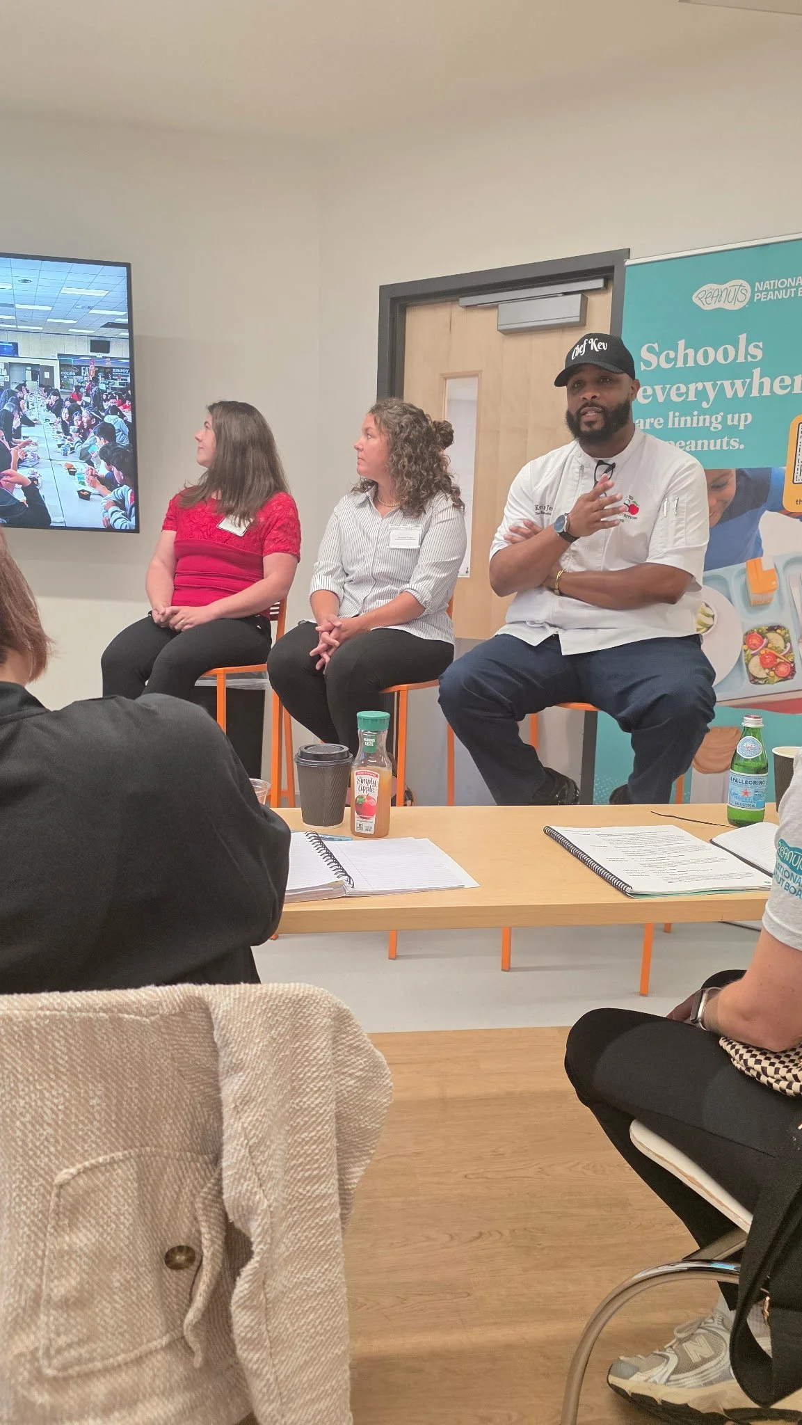 A panel of three speakers, two women and one man, seated on orange chairs at an indoor event, with a large screen displaying an image of children eating at a long table in the background. The man is wearing a black cap and speaking with hands raised, while the women listen attentively.