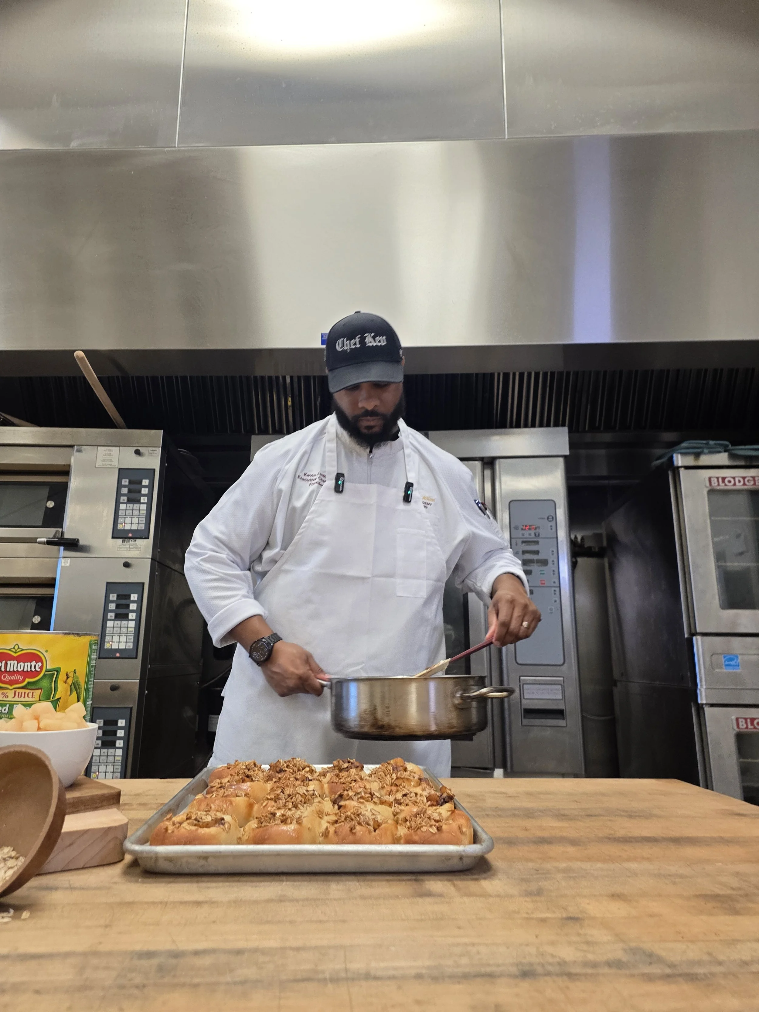 A chef in a kitchen pouring sauce into a baking dish of baked bread rolls topped with crushed nuts, with other baked goods on the counter and kitchen appliances in the background.