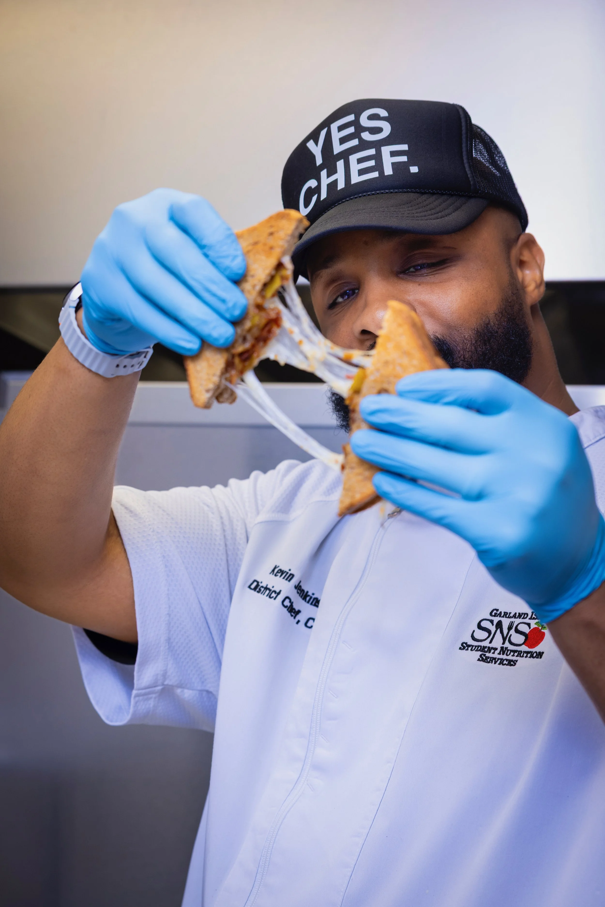 A man wearing a black cap with white lettering that reads "YES CHEF." and a white uniform with a logo and name tag is holding a sandwich with melted cheese and other ingredients, looking at it closely.