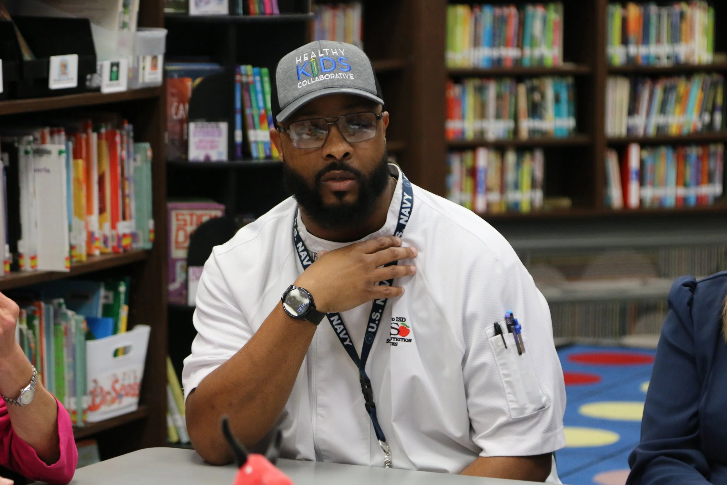 A man with a beard, glasses, and a gray cap that reads "Healthy Kids Collaborative" sits at a table in a library surrounded by colorful bookshelves, wearing a white shirt with a U.S. Navy badge and ID lanyard.