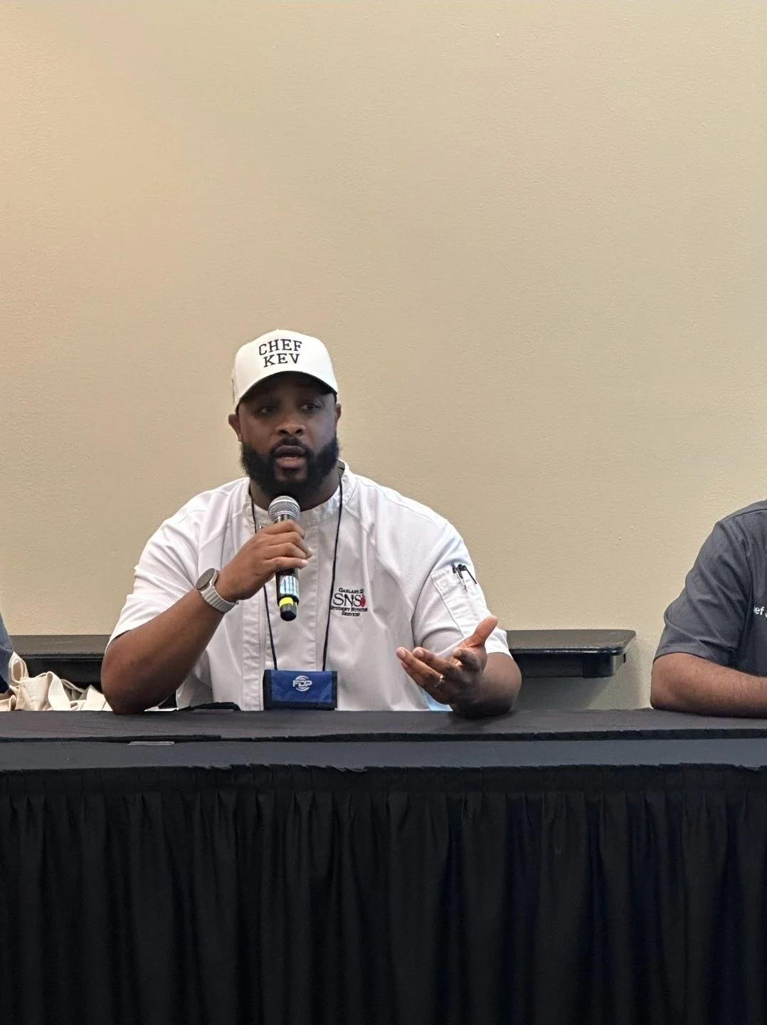 A man in a white chef's coat and a white cap with 'CHEF KEV' written on it, speaking into a microphone at a panel discussion.