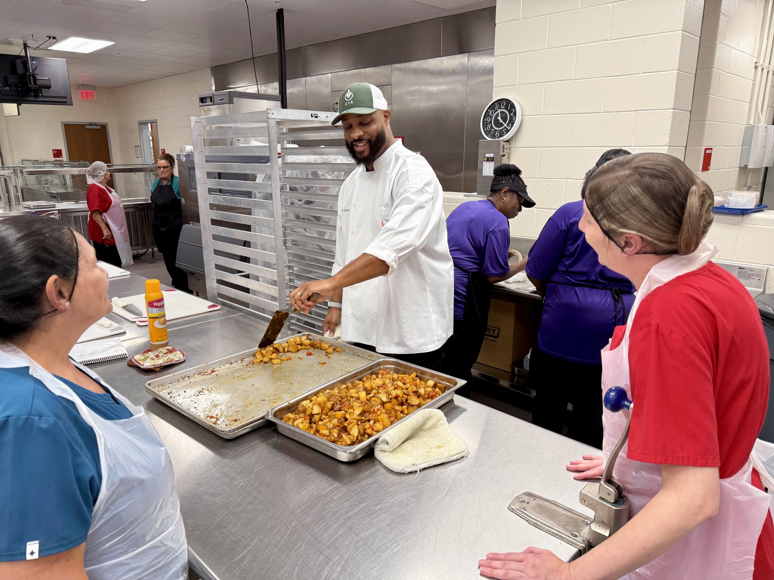 A group of people in a commercial kitchen at a food service or community center, with a chef slicing roasted vegetables on a tray, and others wearing aprons and hair coverings around a stainless steel counter.
