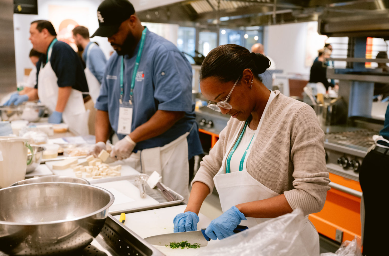 People cooking and chopping vegetables in a kitchen, wearing aprons and gloves, with others preparing food in the background.