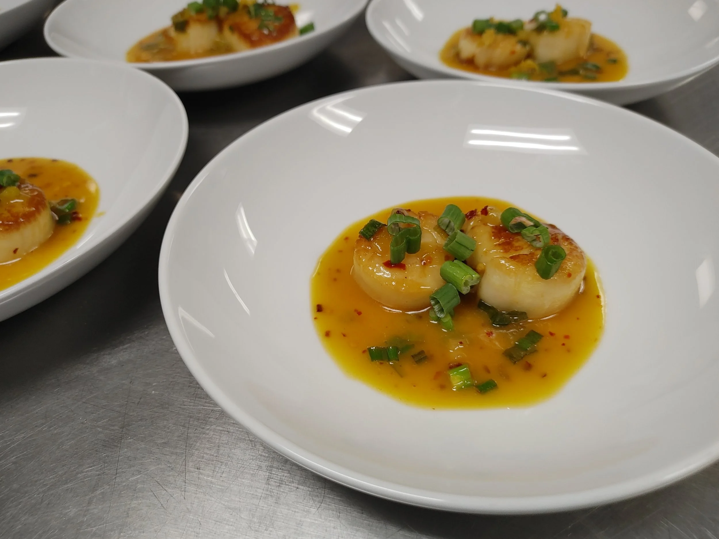 White bowls filled with small, round, seared scallops topped with chopped green onions and drizzled with orange sauce.