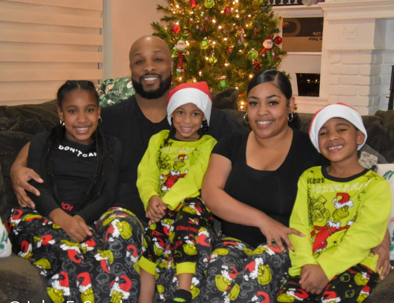 A family sitting on a couch in front of a decorated Christmas tree, all dressed in festive pajamas with holiday characters, some wearing Santa hats, smiling for the camera.