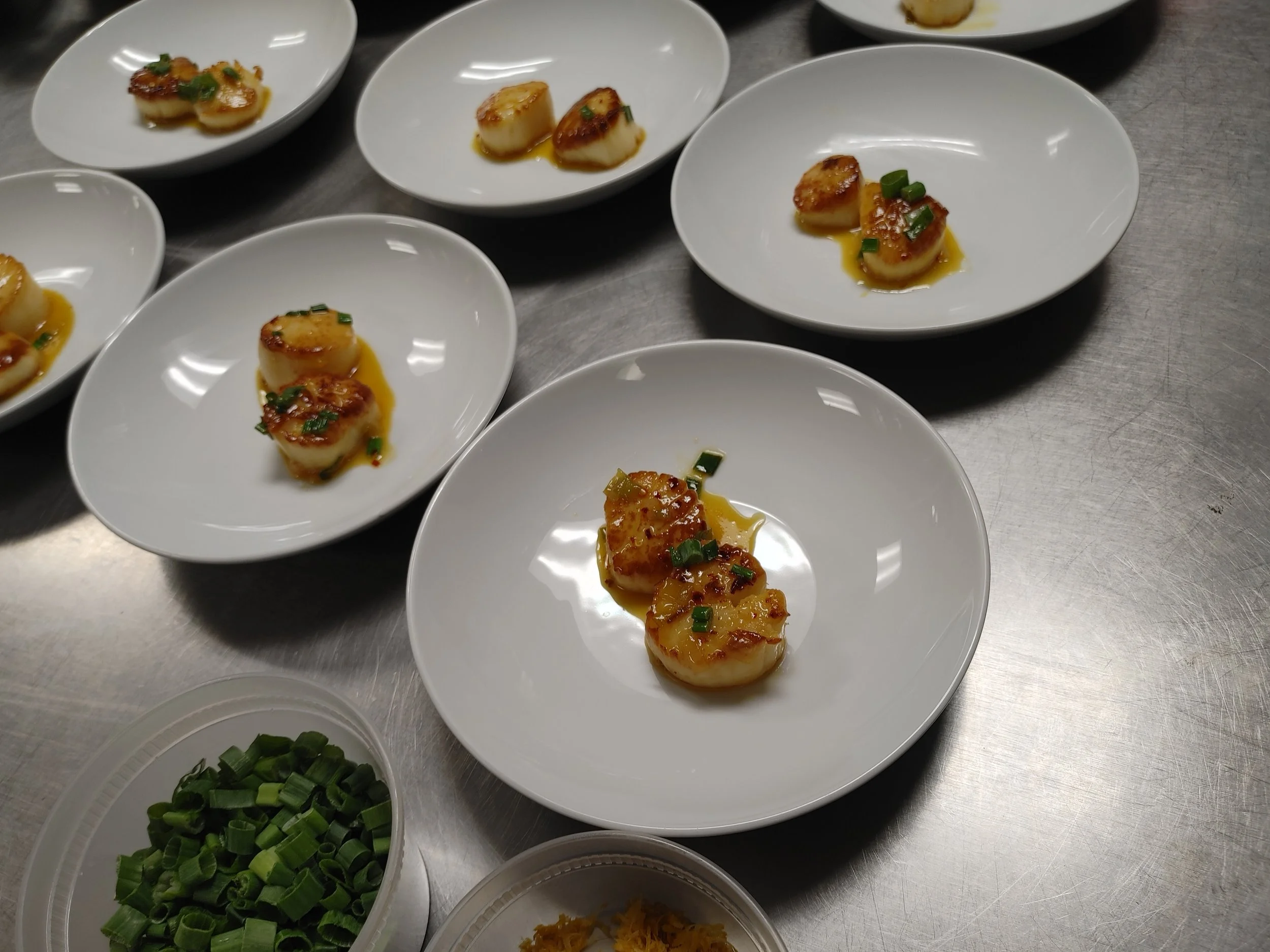 Multiple white plates with scallops garnished with green onions and sauce, and small bowls with chopped green onions, grated cheese, and shredded carrots, on a metallic countertop.