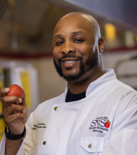 A smiling man in a white chef's coat holding a red apple in a kitchen.