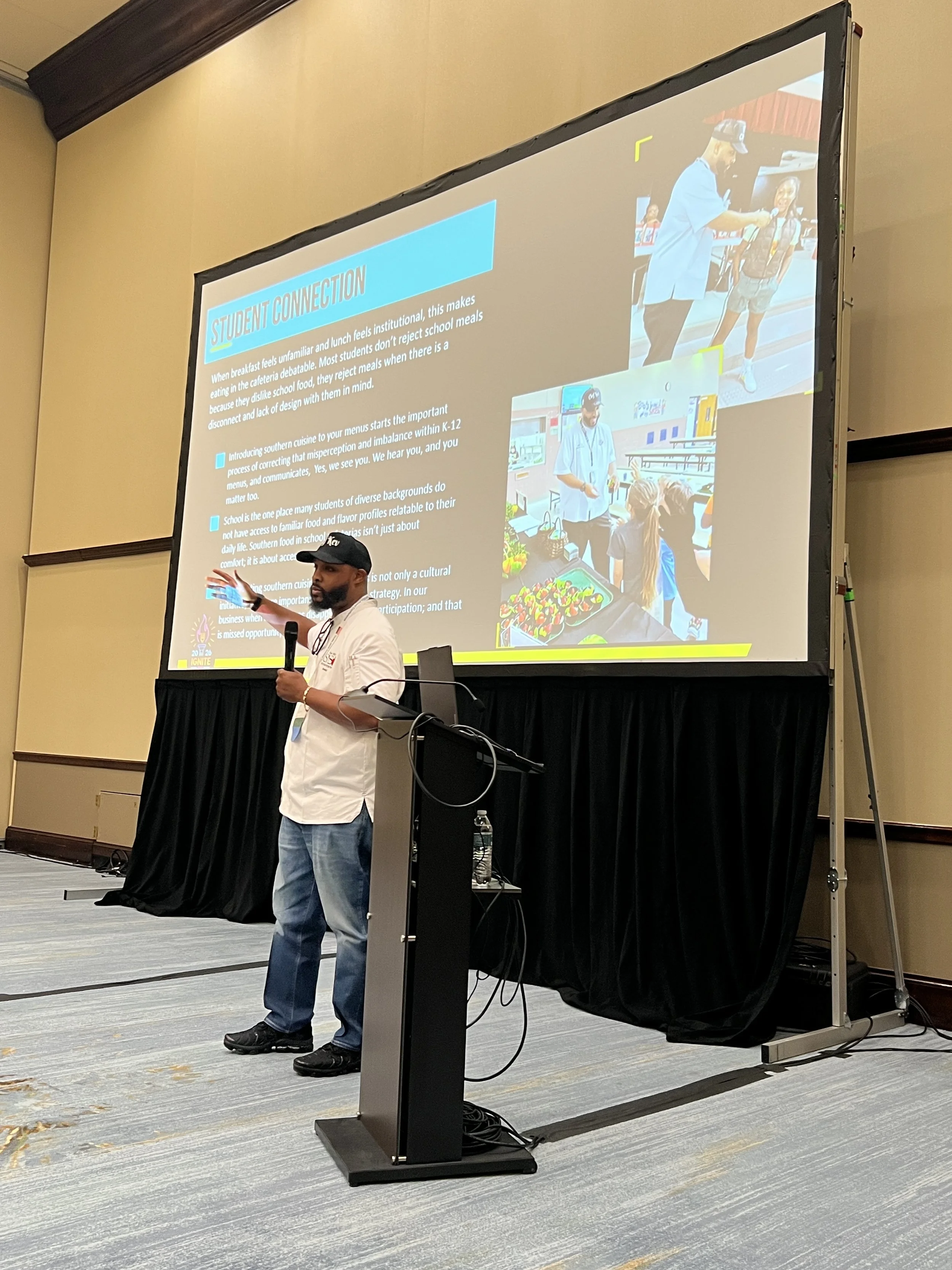 A man with a beard, wearing a black cap, white shirt, and jeans, is giving a presentation in front of a large screen in a conference room. The screen displays a slide titled 'STUDENT CONNECTION' with text and two photos of children at an event.