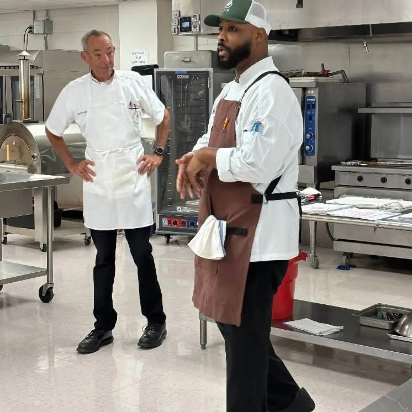 Two chefs in a commercial kitchen having a conversation. One chef is wearing a white uniform and black shoes, the other is wearing a brown apron, black pants, and a green and white cap.