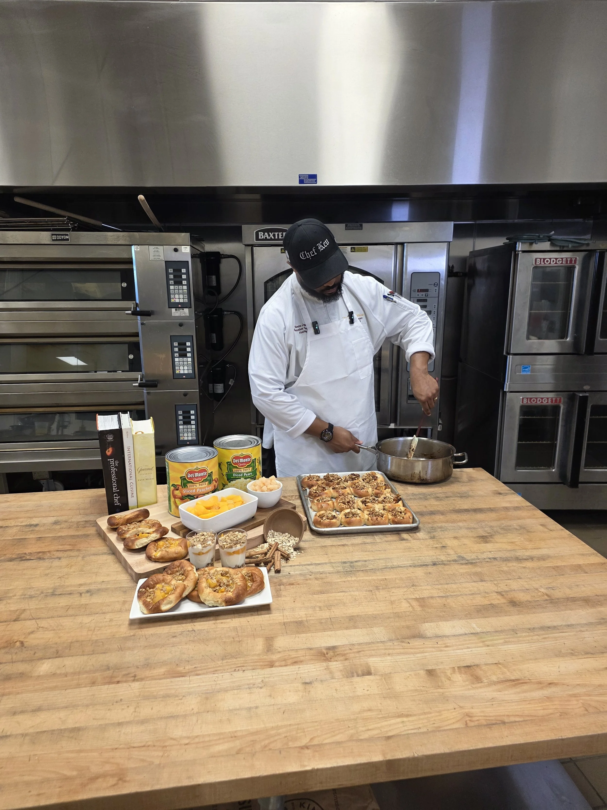 A chef wearing a black cap that reads 'Chef Ale' and a white apron is cooking in a commercial kitchen. The counter in front of him is filled with baked goods such as bread rolls, danishes, and pastries, along with canned peaches, a bowl of diced peac