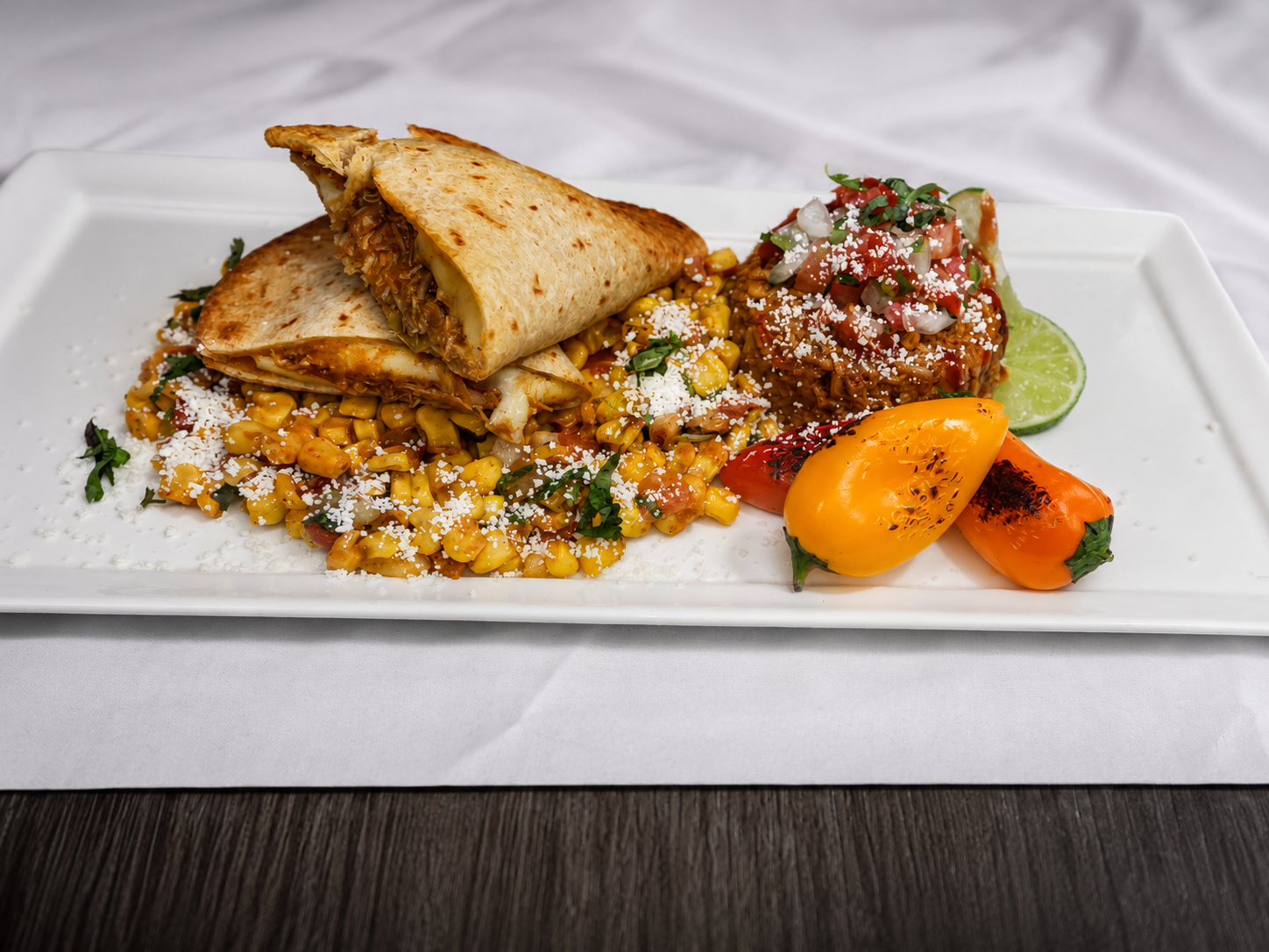 Plate with grilled yellow corn salsa, a folded grilled quesadilla, chopped tomato and onion salad with cheese, lime wedge, and grilled yellow and orange peppers on a white rectangular plate.
