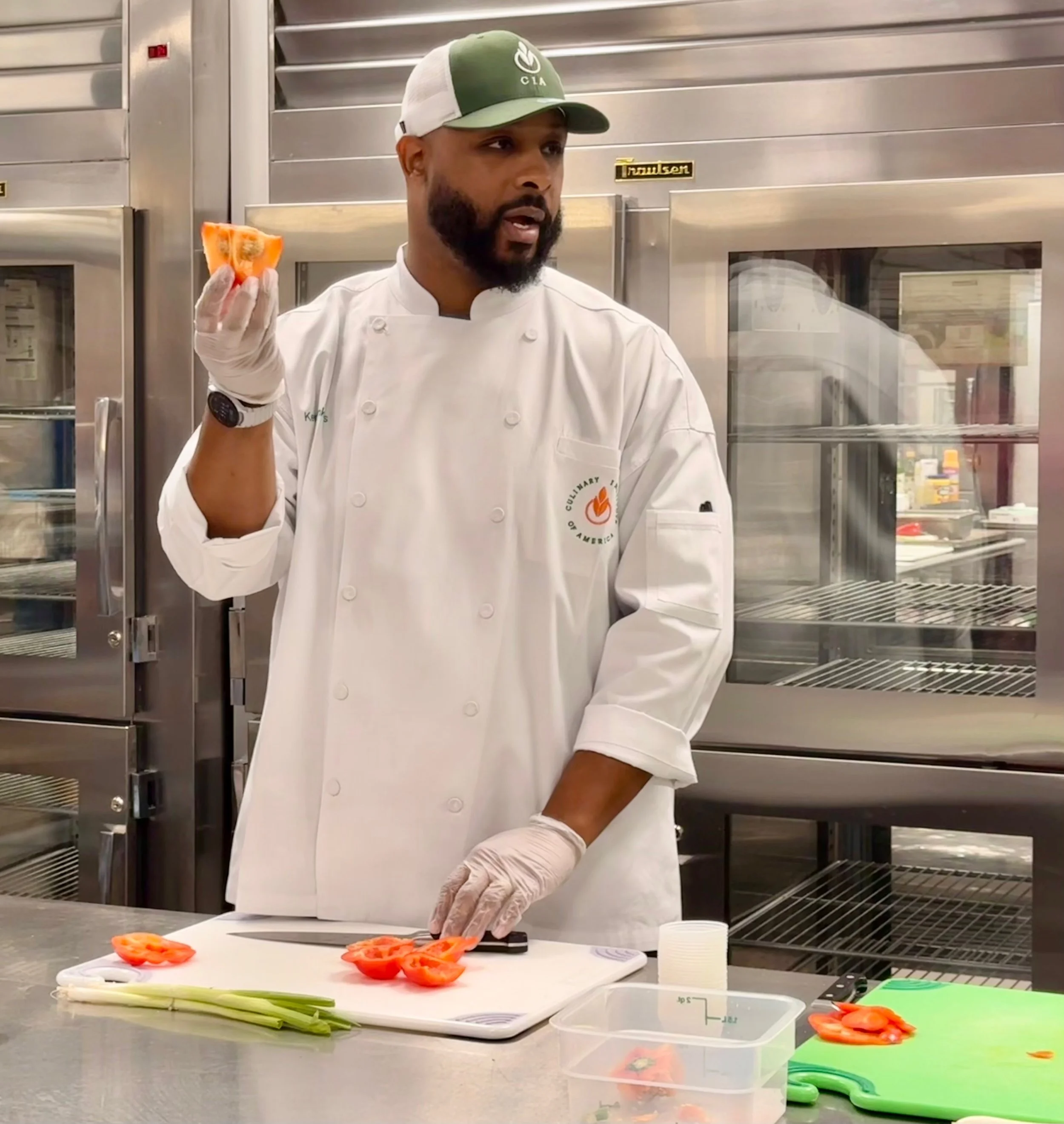 A chef in a white uniform and green cap holding a sliced orange bell pepper while standing in a commercial kitchen. There are sliced bell peppers, green onions, and kitchen tools on the stainless steel counter, with a large stainless steel refrigerat