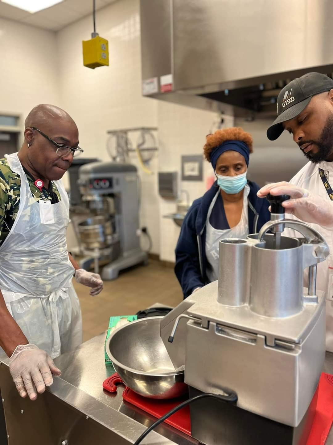 Three people in a kitchen, two men and one woman, are wearing aprons and gloves while observing a machine. One woman is wearing a face mask. The kitchen has industrial equipment in the background.