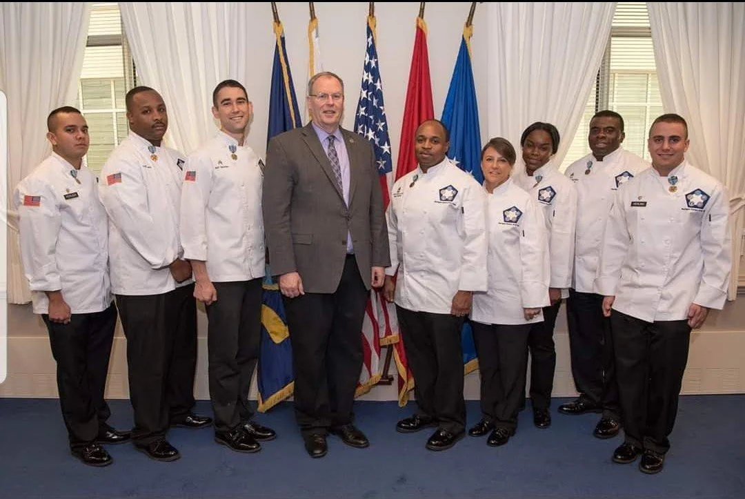 Group of nine people, including seven in uniform and two in suits, standing indoors in front of flags, smiling for a photo.