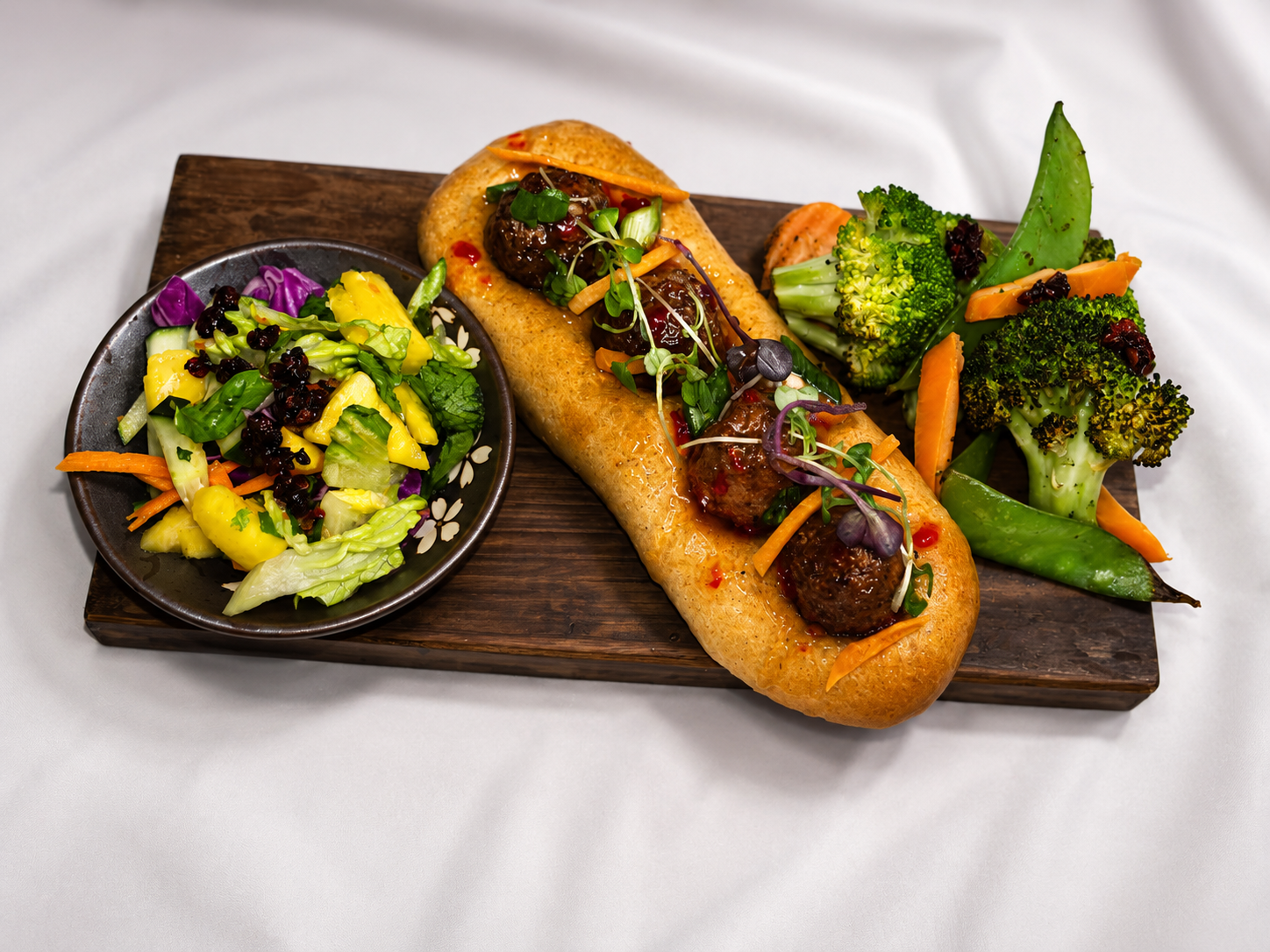 A wooden serving board with a sandwich containing meatballs, a side salad with mixed greens, purple cabbage, and yellow fruit, and a variety of steamed vegetables including broccoli, carrots, snap peas, and snow peas.