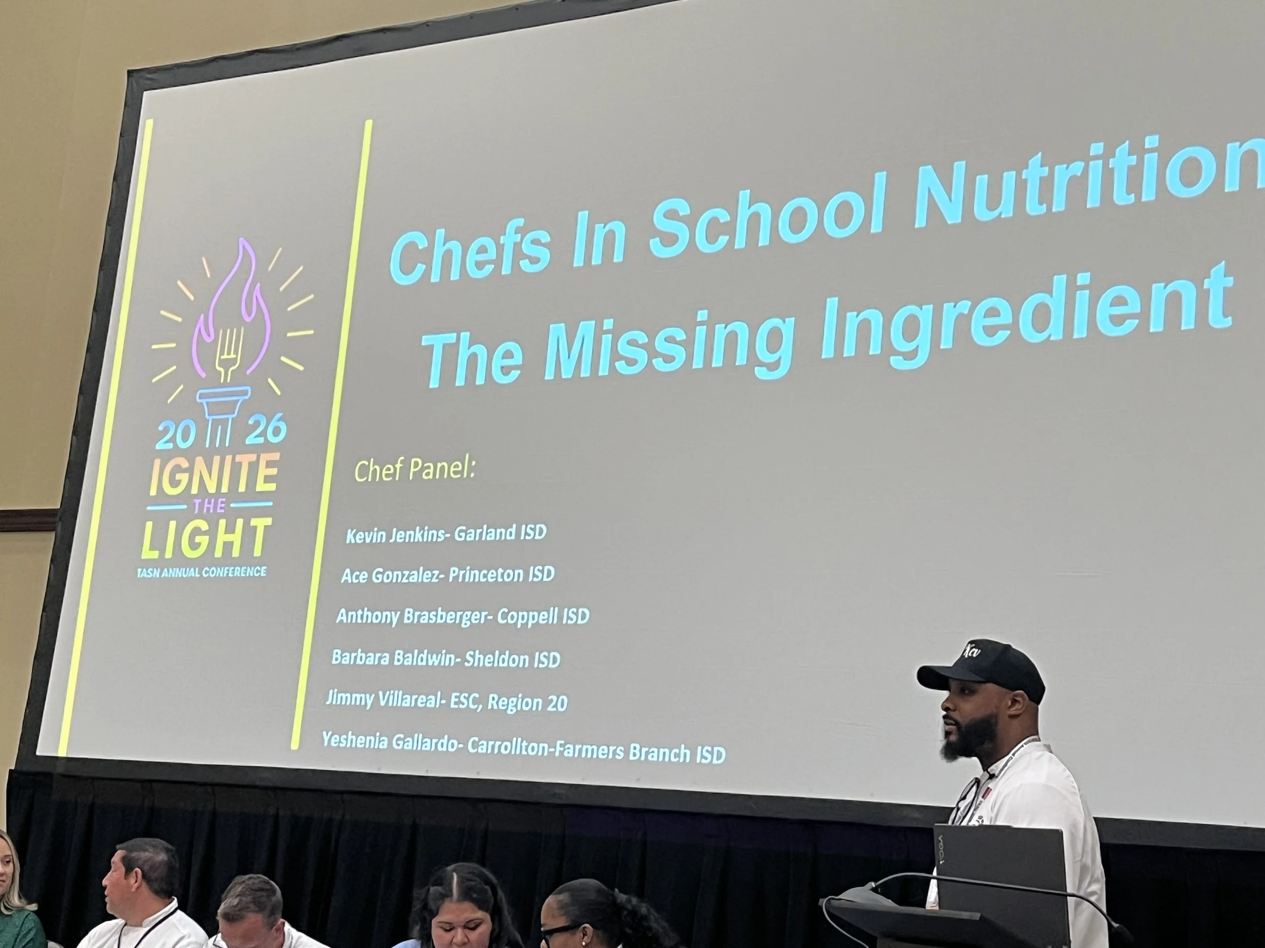 A man in a baseball cap and white shirt standing in front of a large screen during a presentation at a conference. The screen displays the title "Chefs In School Nutrition: The Missing Ingredient" and a list of speakers' names and school districts.