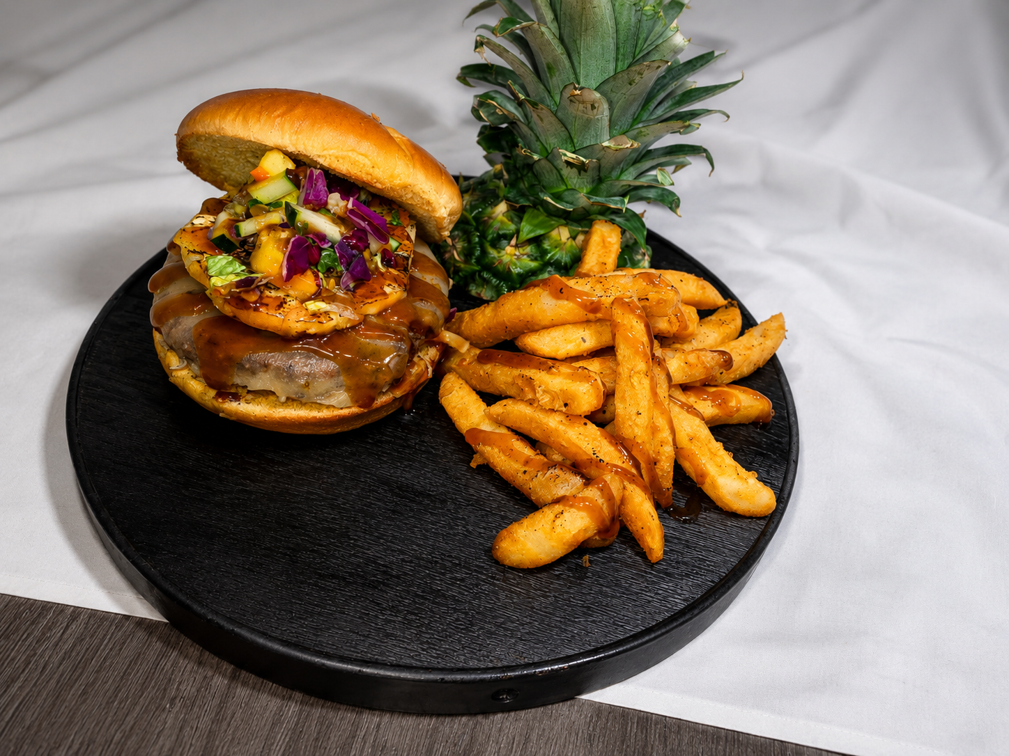 A black plate with a burger topped with various vegetables, barbecue sauce, and a toasted bun, along with crispy French fries drizzled with sauce and a fresh pineapple on a white tablecloth.