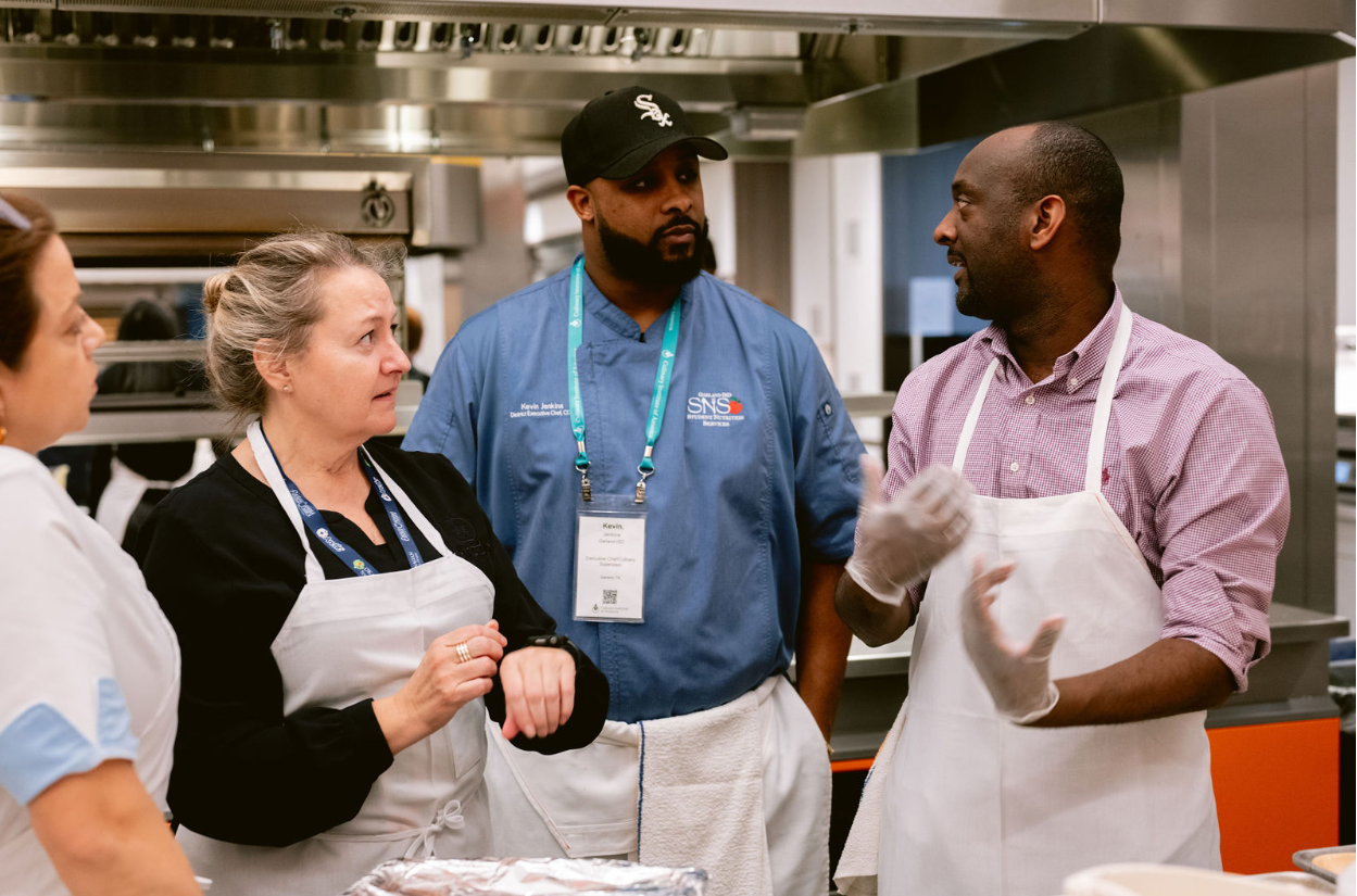 Four people, including a chef and three women, are having a discussion in a professional kitchen. The chef and one woman are wearing aprons, while another woman is listening attentively. The chef appears to be explaining something.