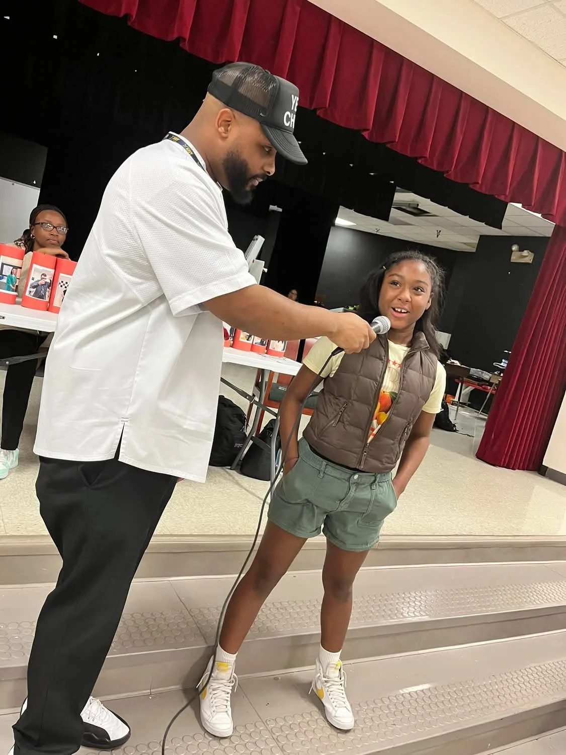 A man in a white shirt and black cap is helping a young girl speak into a microphone on a stage with red curtains. A girl in the background is sitting at a table observing.