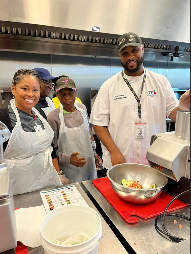 Group of four diverse people in a kitchen, wearing aprons. One man in a chef's coat and hat is mixing ingredients in a large bowl, smiling at the camera. The others watch him, some smiling, one giving a thumbs up.
