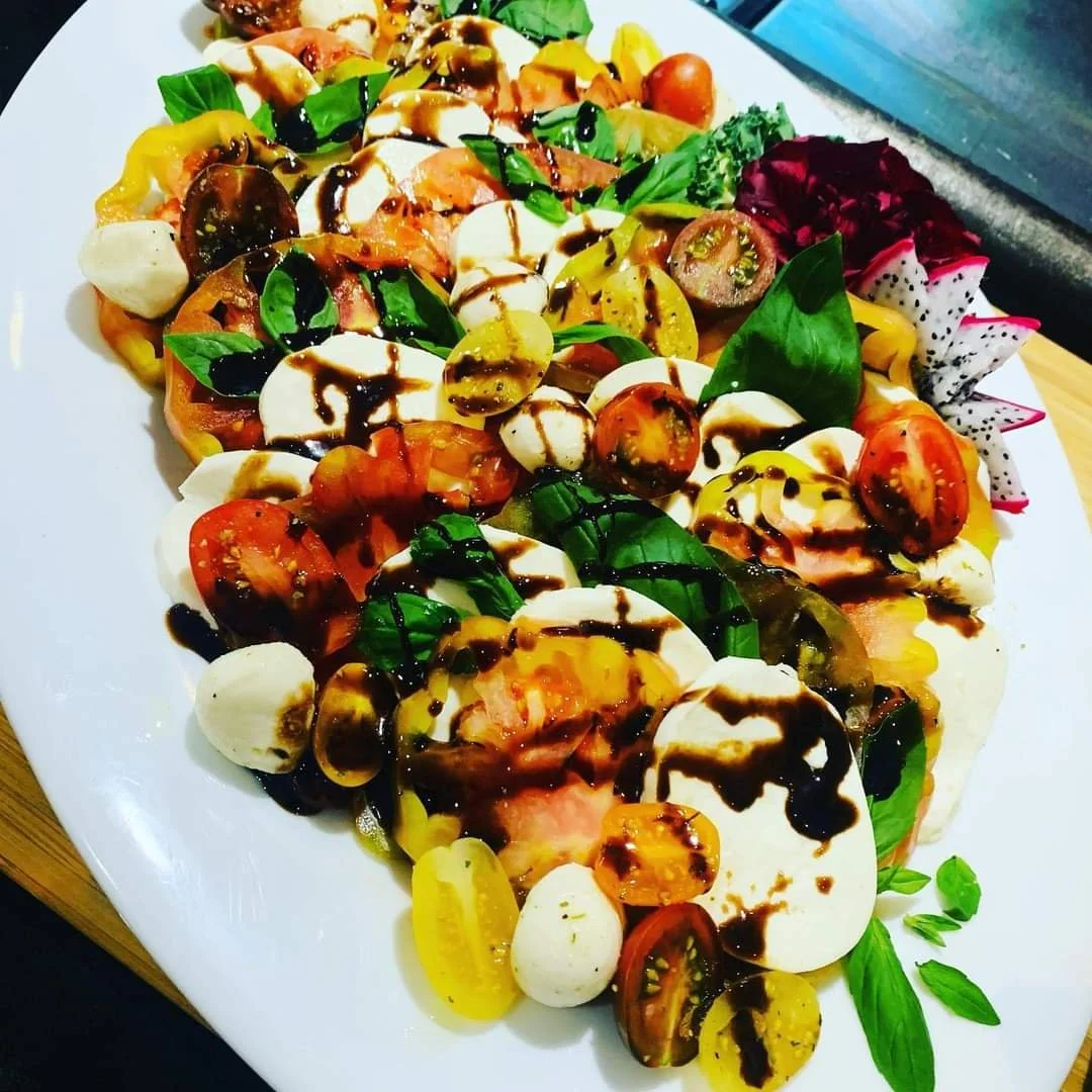 A colorful Caprese salad with fresh cherry tomatoes, mozzarella balls, basil leaves, drizzle of balsamic vinegar and olive oil on a white plate.
