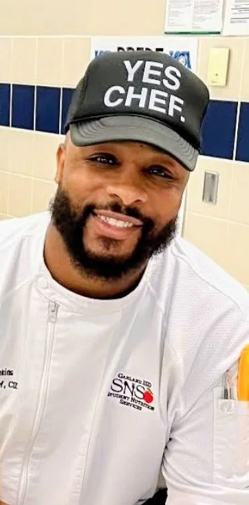 A smiling man wearing a black cap that says 'YES CHEF.' and a white chef's jacket in a kitchen setting.