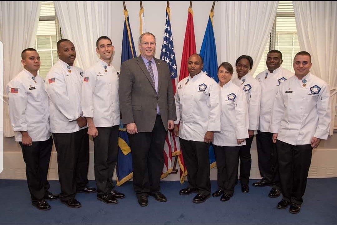 Group of nine people, including eight men and one woman, standing in front of a row of flags inside a room with white curtains.