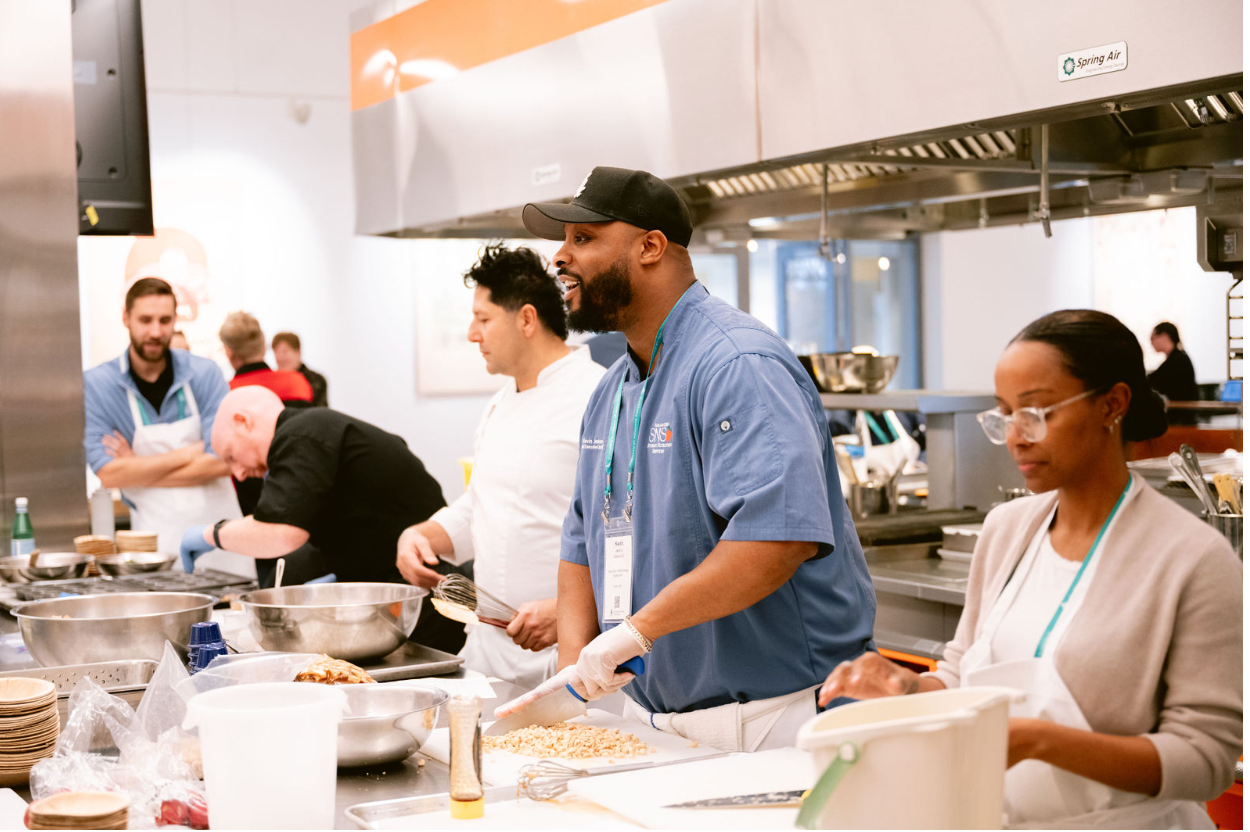 A group of four people in a professional kitchen, preparing food. The man in the foreground wears a blue shirt, black cap, and gloves, and is chopping nuts. Next to him, a woman in glasses and a beige sweater looks down at her work. Behind them, two men, one in a chef's coat and the other in casual attire, are also working. There is a stainless steel stove, kitchen utensils, and ingredients on the counters.