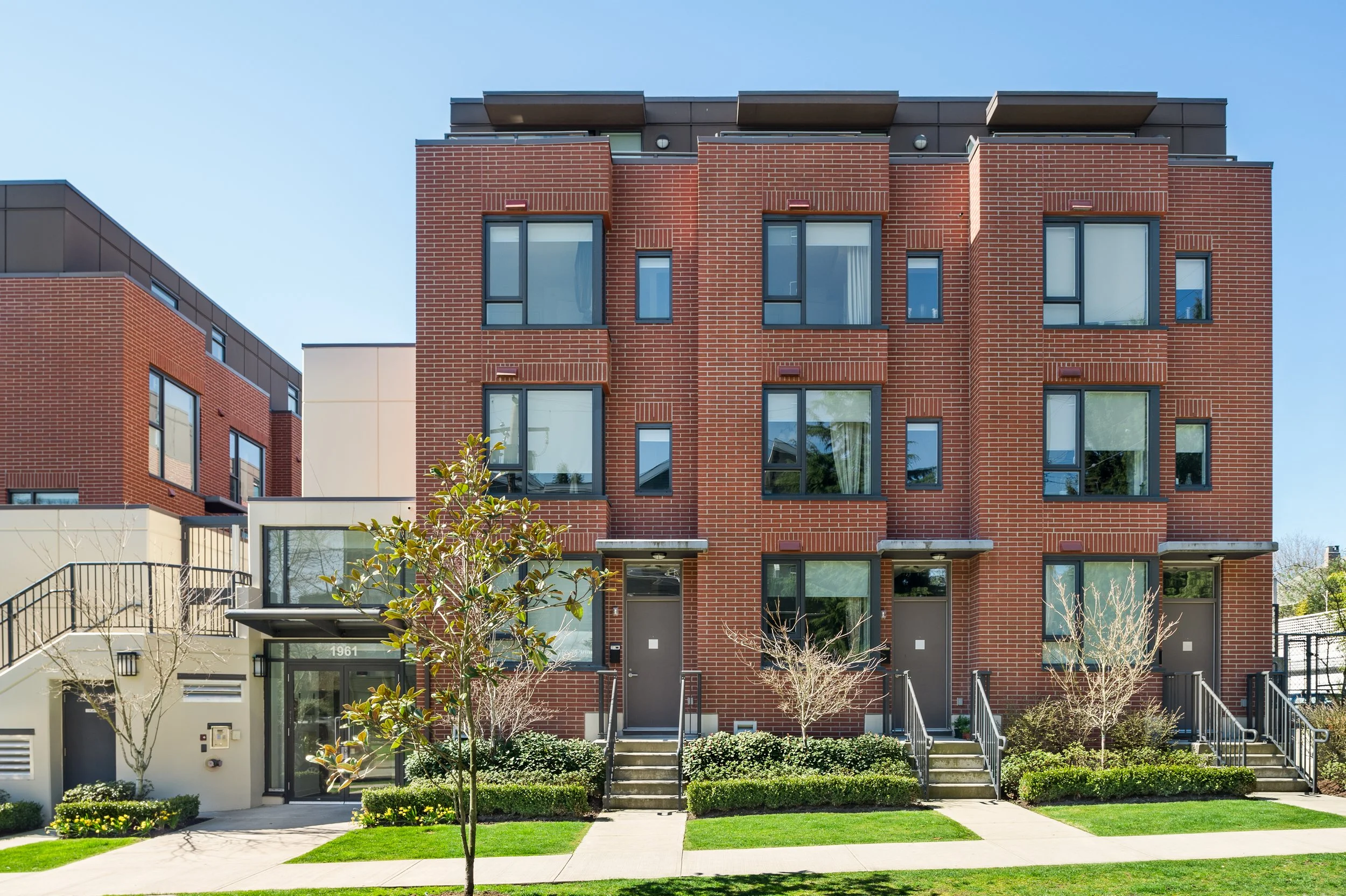Front view of modern multi-story residential building with brick exterior, large windows, small trees and landscaping in front, clear blue sky.