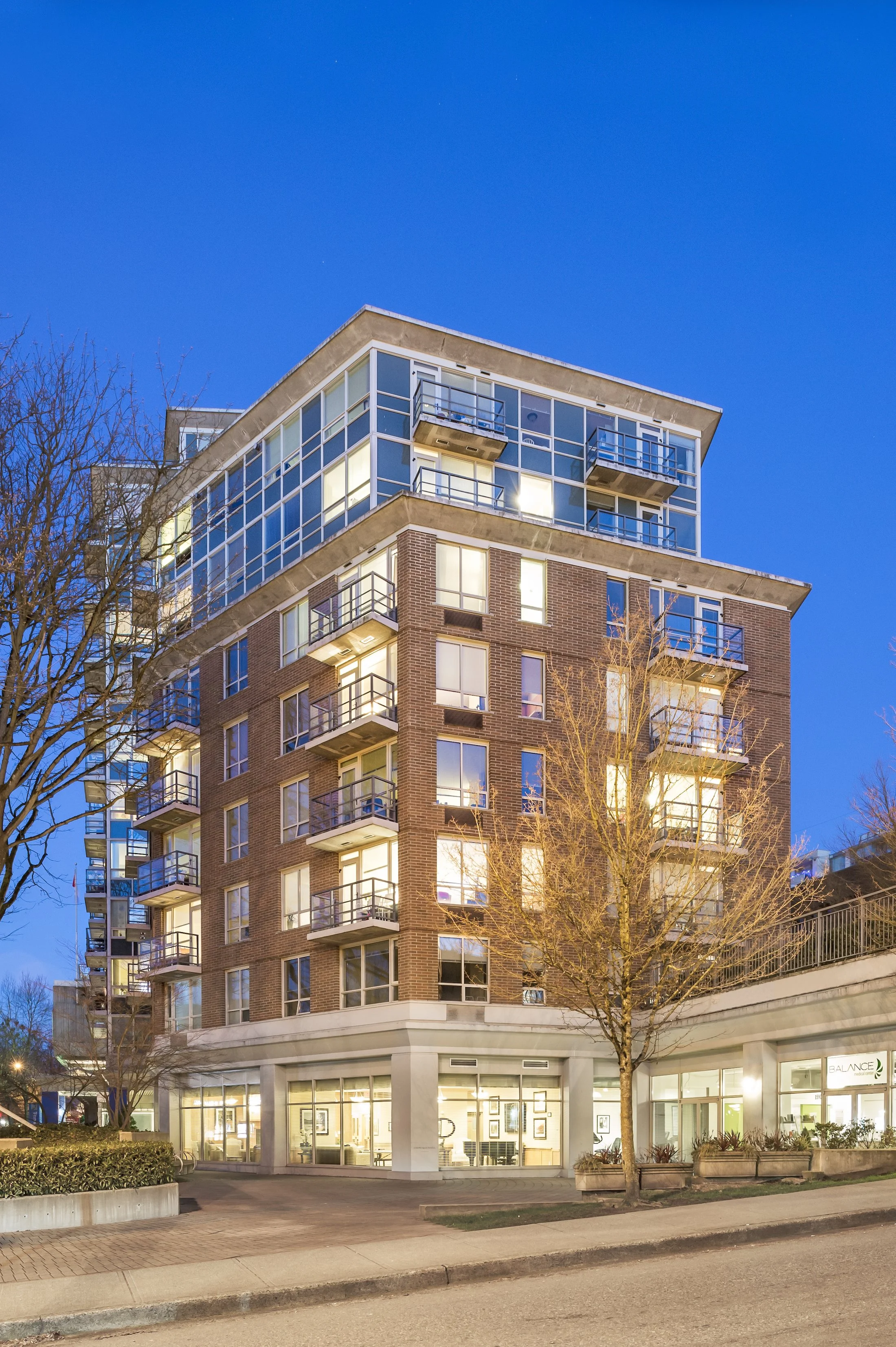 A multi-story residential building with glass balconies and brick exterior, illuminated from within during dusk. The ground level features a commercial space with large windows and a sign that reads 'BALANCE,' possibly a wellness or fitness center. There are leafless trees and a sidewalk in front of the building.