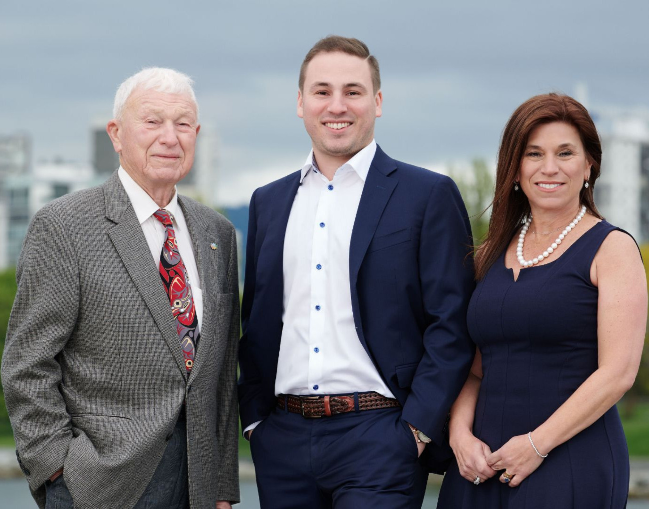 Three people standing outdoors in front of city buildings, smiling. An elderly man on the left wearing a gray plaid suit and red patterned tie, a young man in the middle wearing a navy suit, white shirt, and brown belt, and a woman on the right with shoulder-length brown hair wearing a navy dress and pearl necklace.