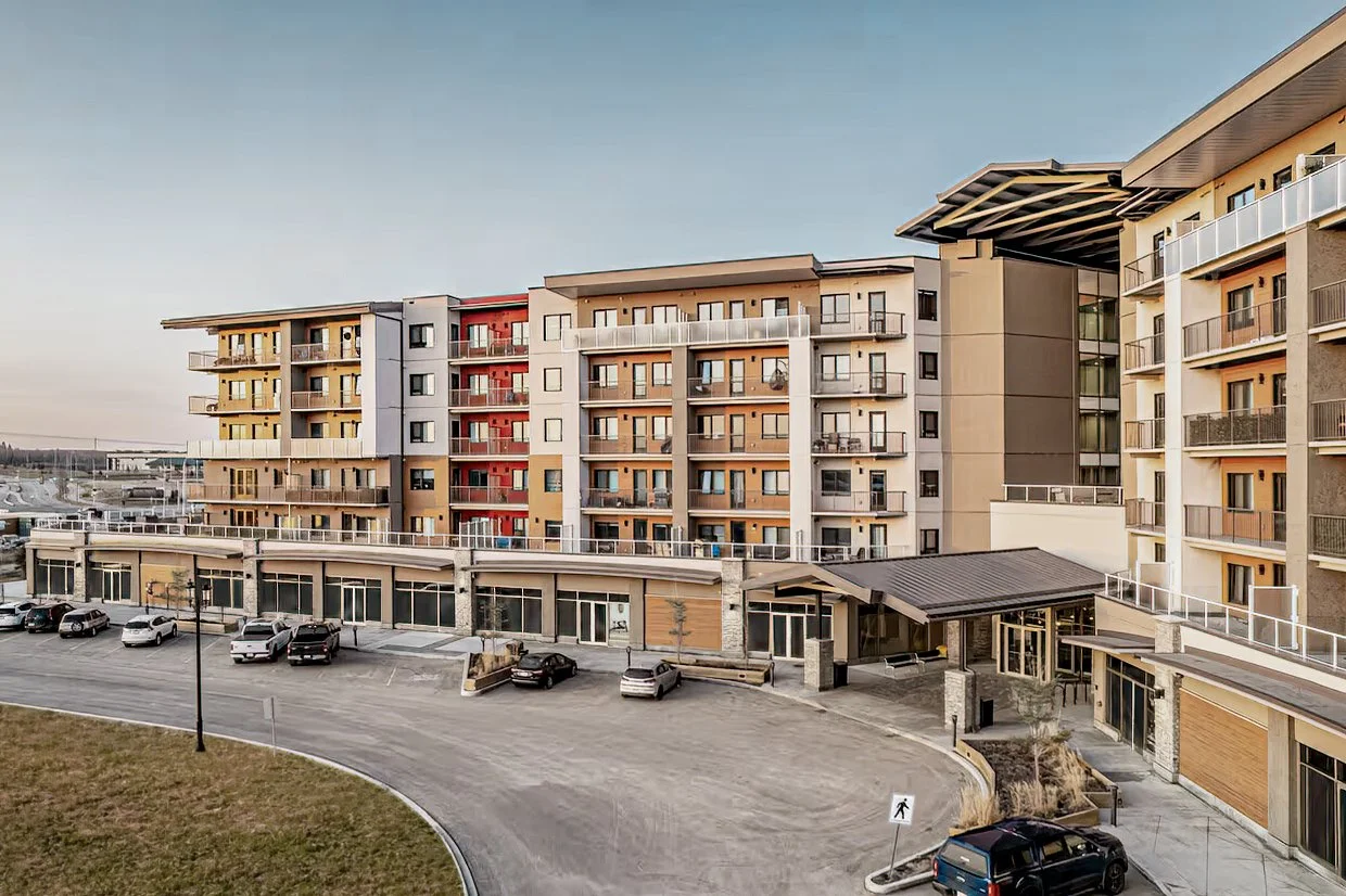 Multi-story modern apartment building with balconies, parking lot, and landscaped area in front.