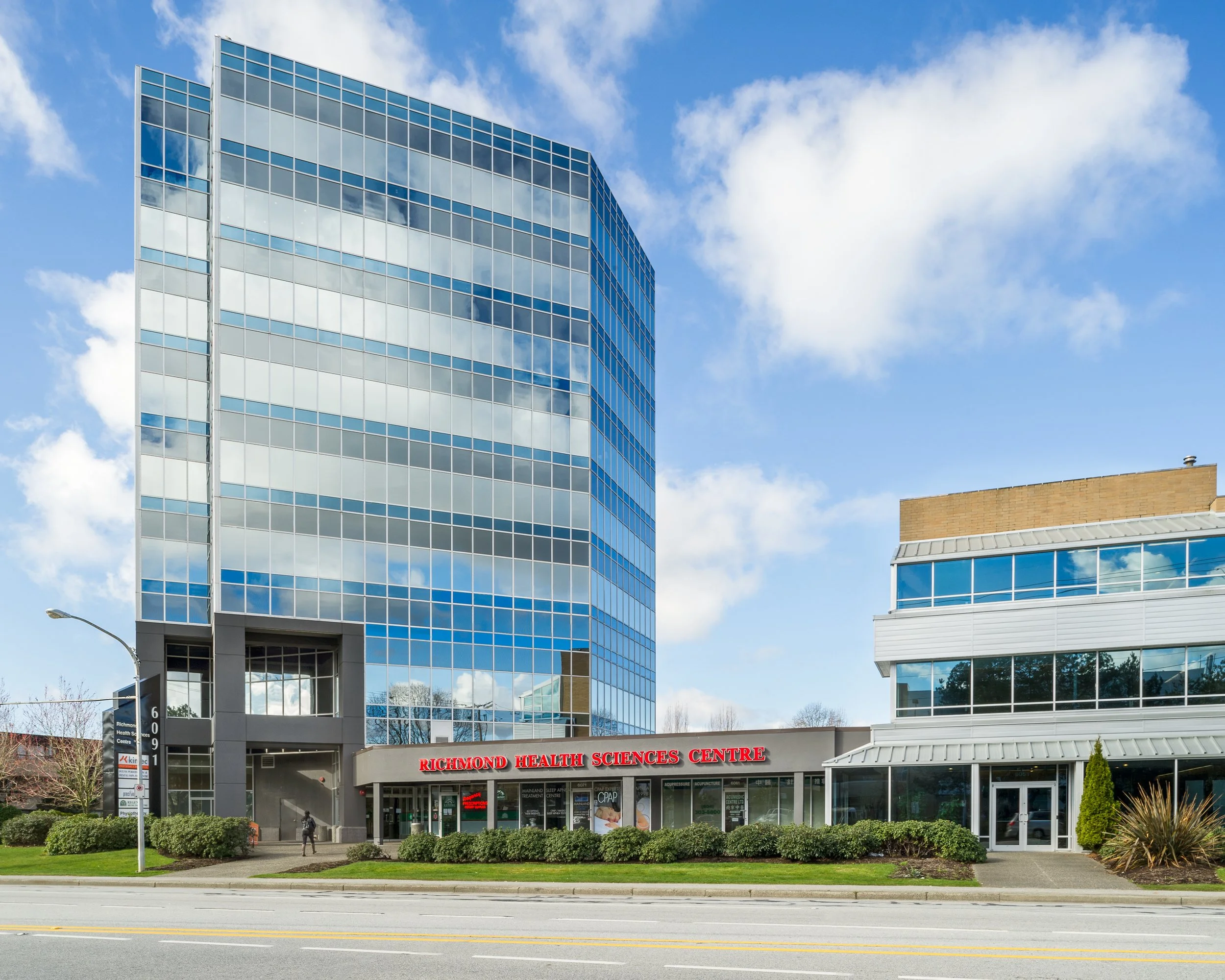 A modern multi-story office building with glass windows reflecting clouds and sky, and a sign for Richmond Health Sciences Centre at the entrance, with a clear blue sky overhead.