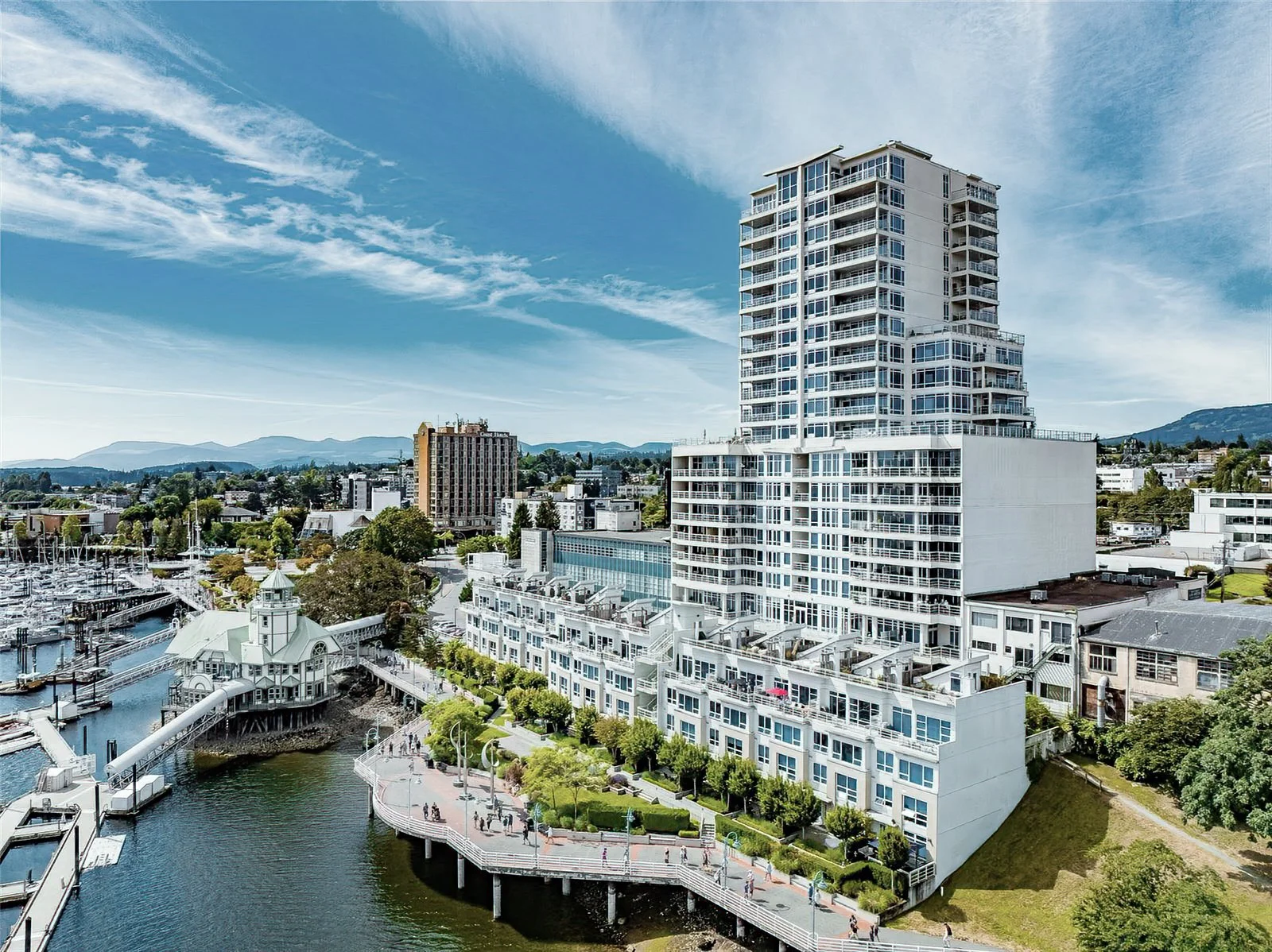 A cityscape featuring a tall modern white building near a body of water, with a marina and sailboats, and a boardwalk with people walking. In the background, there are other buildings, green trees, and distant mountains under a partly cloudy blue sky.