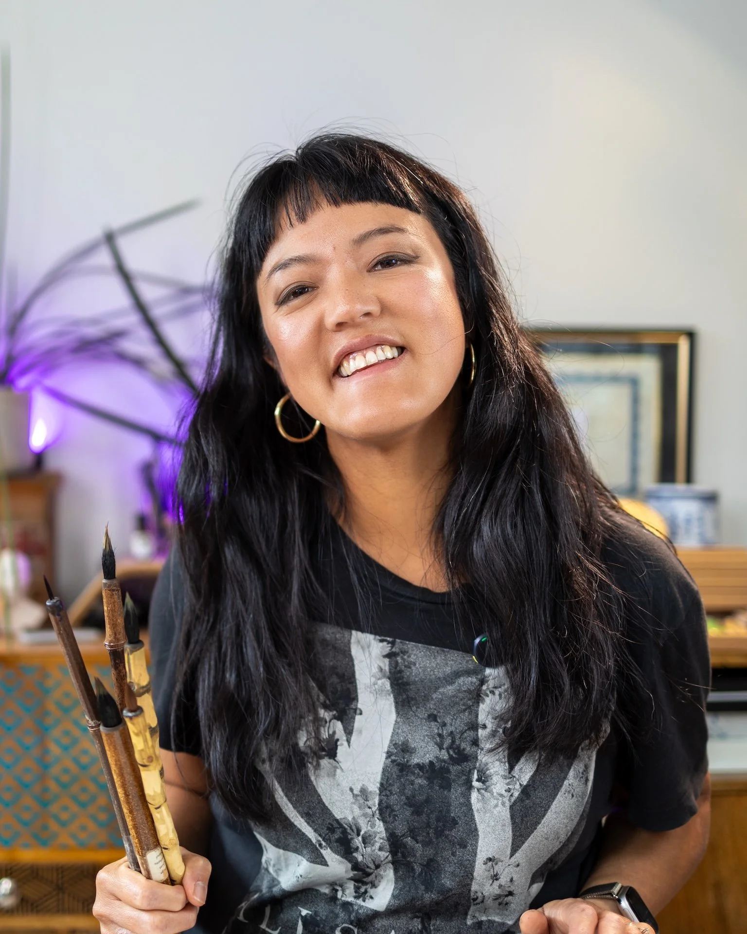 A woman with black hair and a fringe, wearing hoop earrings, holding traditional Chinese calligraphy brushes, smiling in a creative workspace.