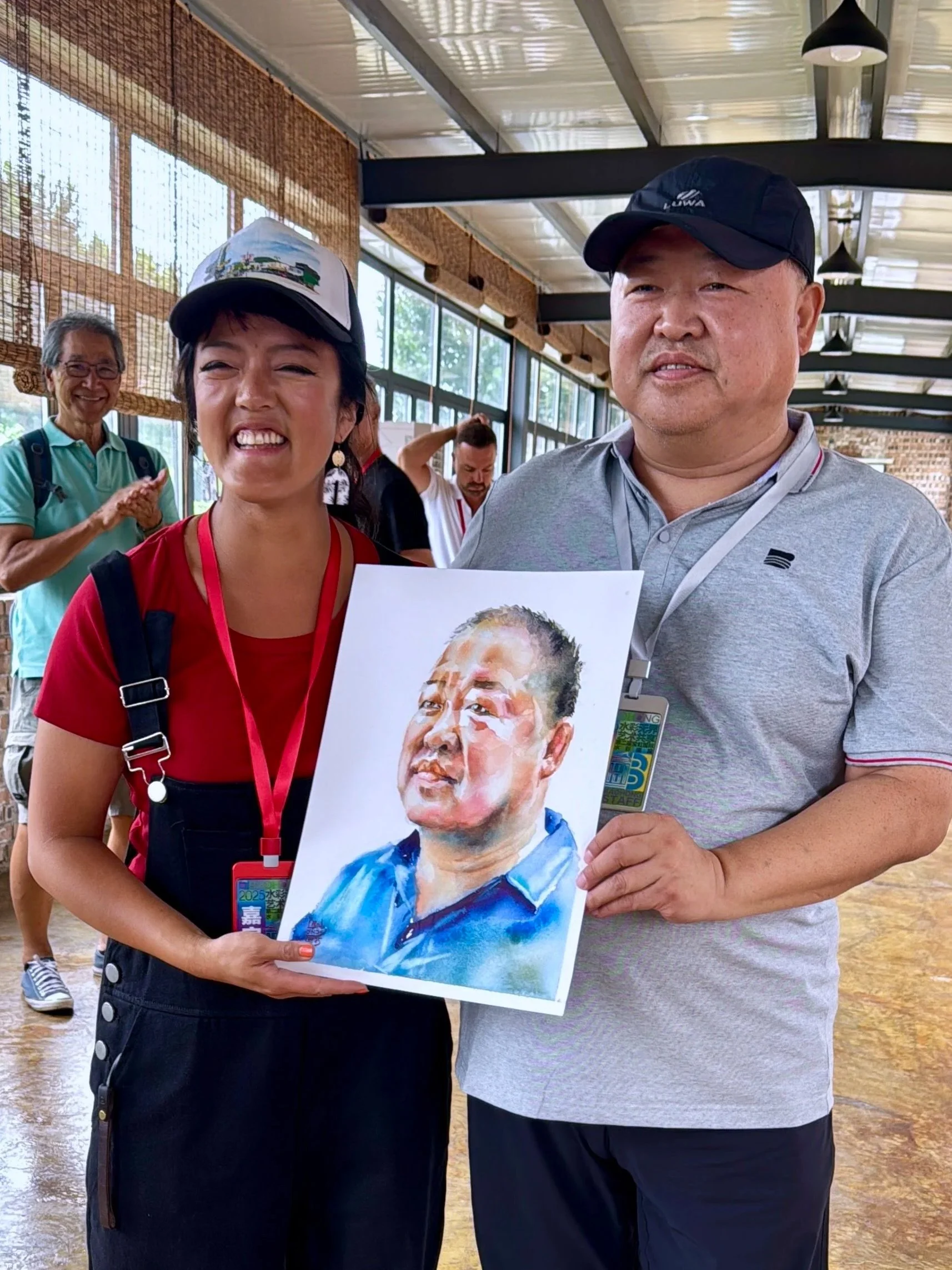 Two people smiling at an indoor event, with one holding a watercolor portrait of a man wearing a blue shirt. Several other people are in the background, some clapping and one adjusting his hair. The setting has a high ceiling with industrial-style li