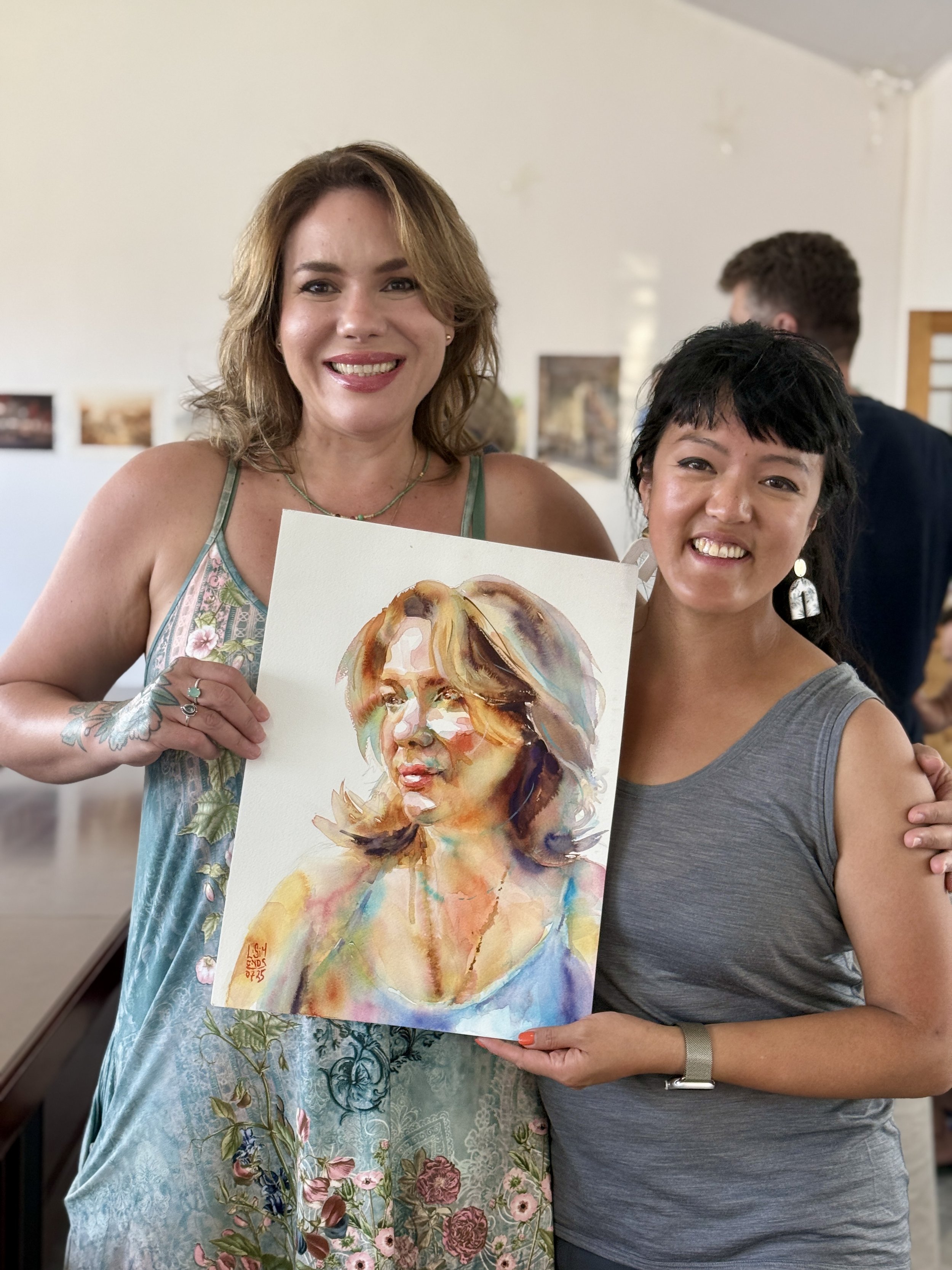 Two women smiling and holding a watercolor painting of a woman's portrait. The woman on the left has light brown hair, a sleeveless floral dress, and is wearing rings and a necklace. The woman on the right has dark hair, a gray tank top, earrings, an