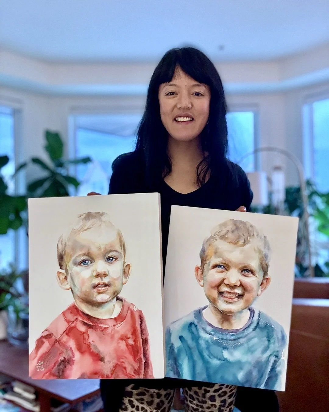 Lorraine Simonds holding two watercolor portraits of young boys, one with a serious expression in a red shirt, and the other smiling in a blue shirt, in a well-lit room with large windows and green plants in the background.