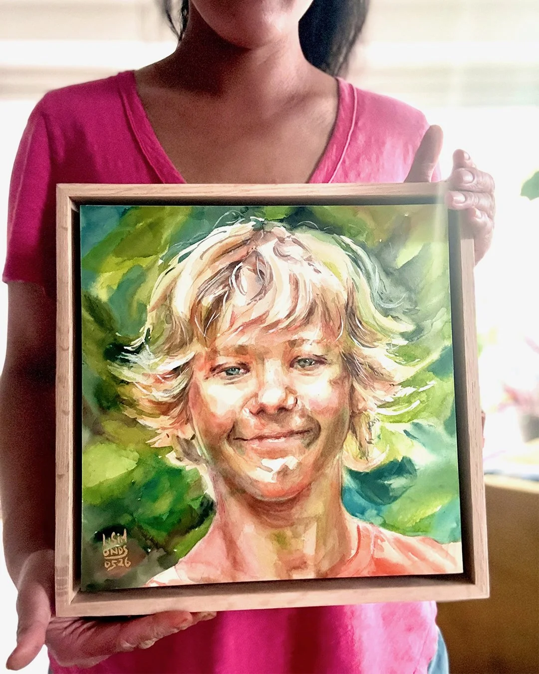 A woman in a pink shirt is holding a framed watercolor portrait of a smiling woman with short blonde hair, set against a green leafy background.