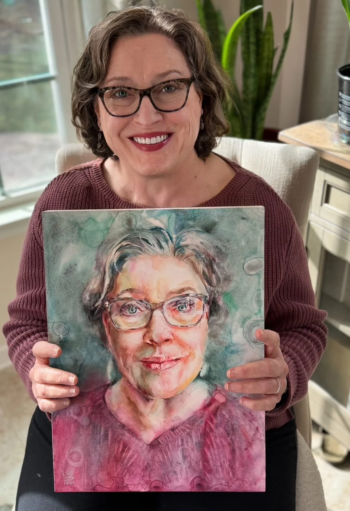 A woman with glasses and short curly hair smiling while holding a watercolor portrait of herself that resembles her.