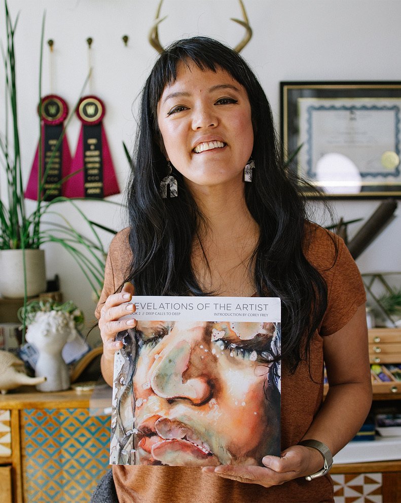 A woman with long black hair, wearing a brown shirt, smiling, and holding an art book titled 'Revelations of the Artist' featuring a watercolor portrait of a person's face.