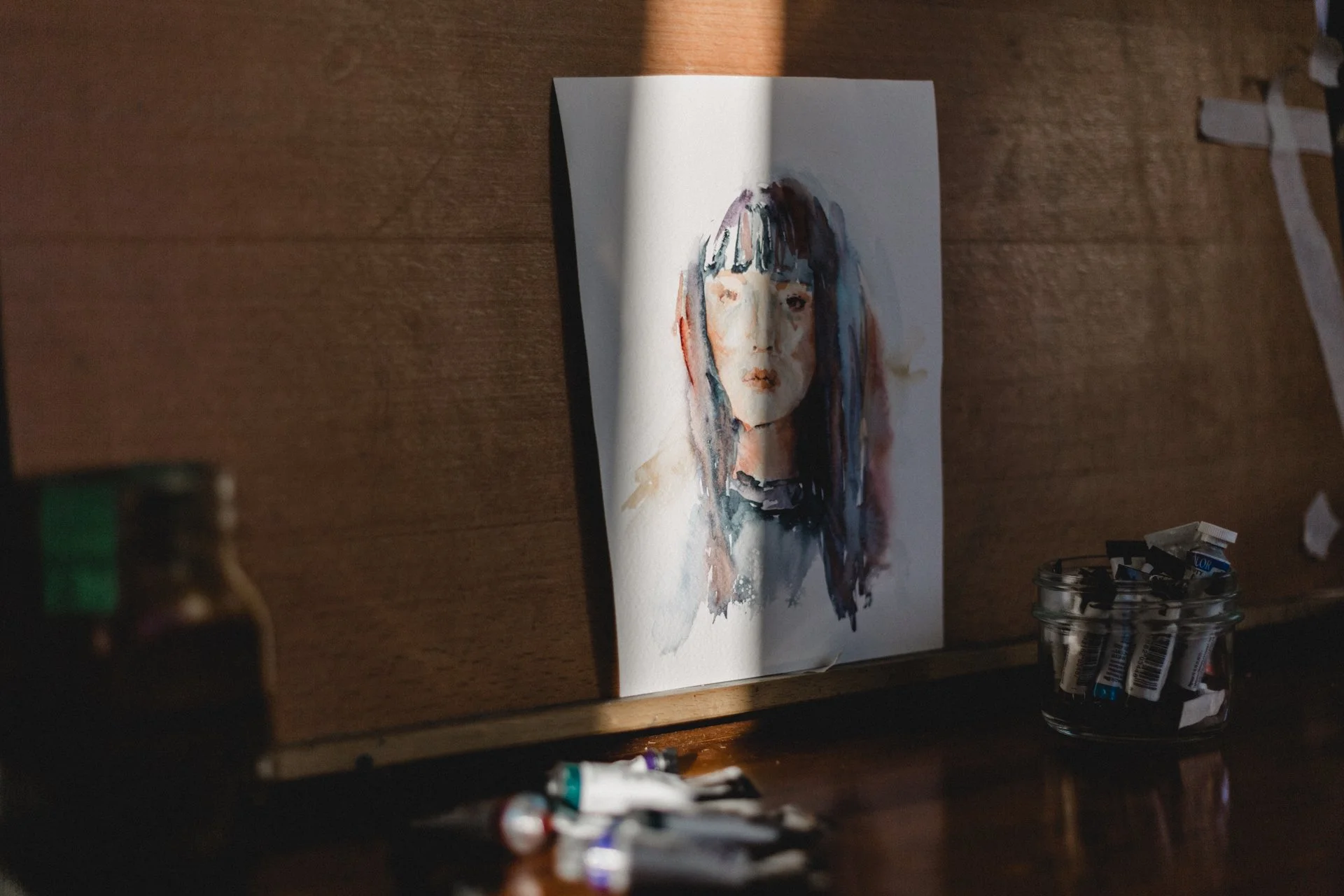 Watercolor portrait of a woman with dark hair and bangs on a wooden surface, illuminated by sunlight, beside small paint tubes and a jar of brushes.