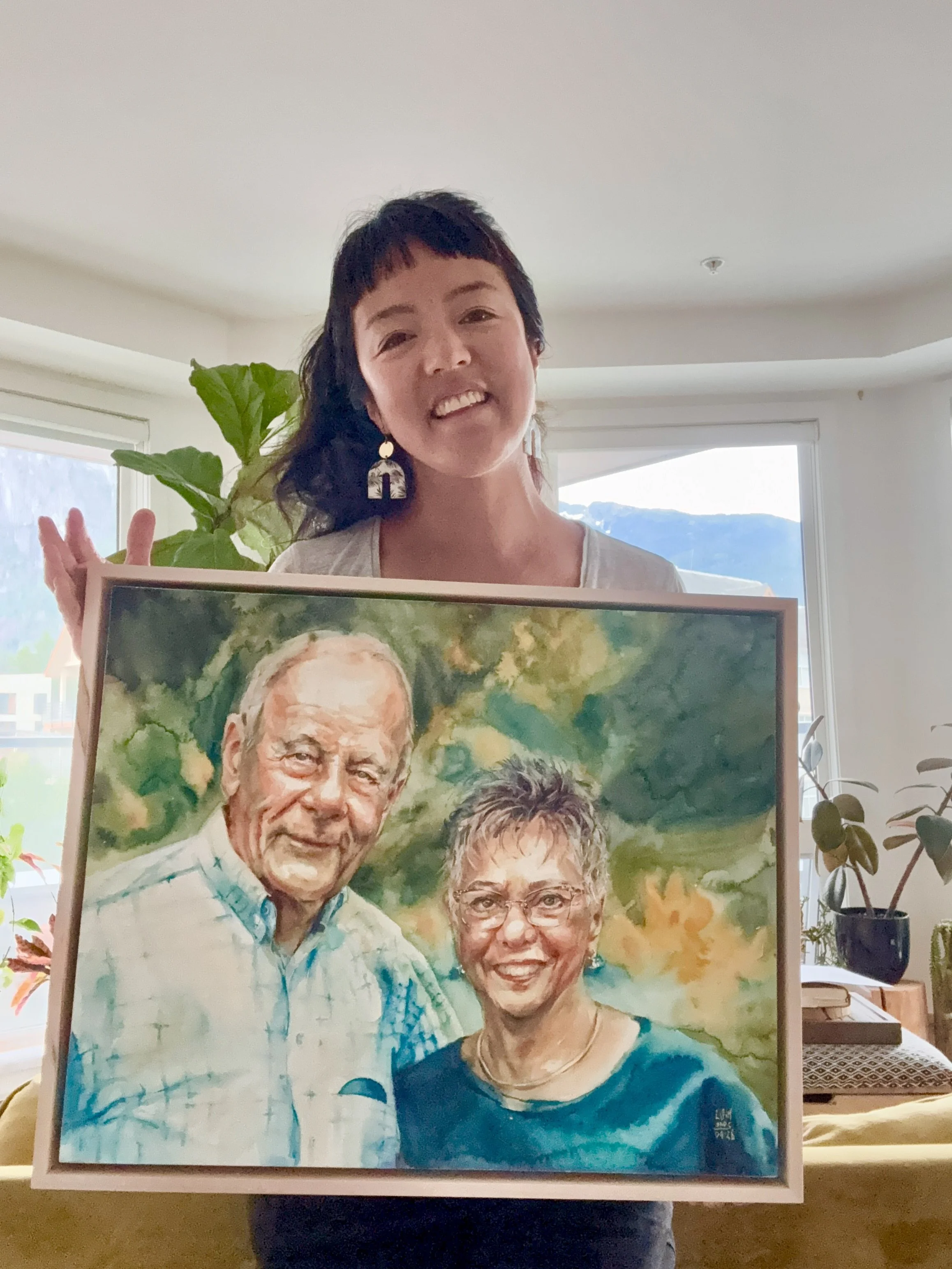Lorraine Simonds smiling and holding a framed painting of four older adults, with a mountain visible through a window in the background.