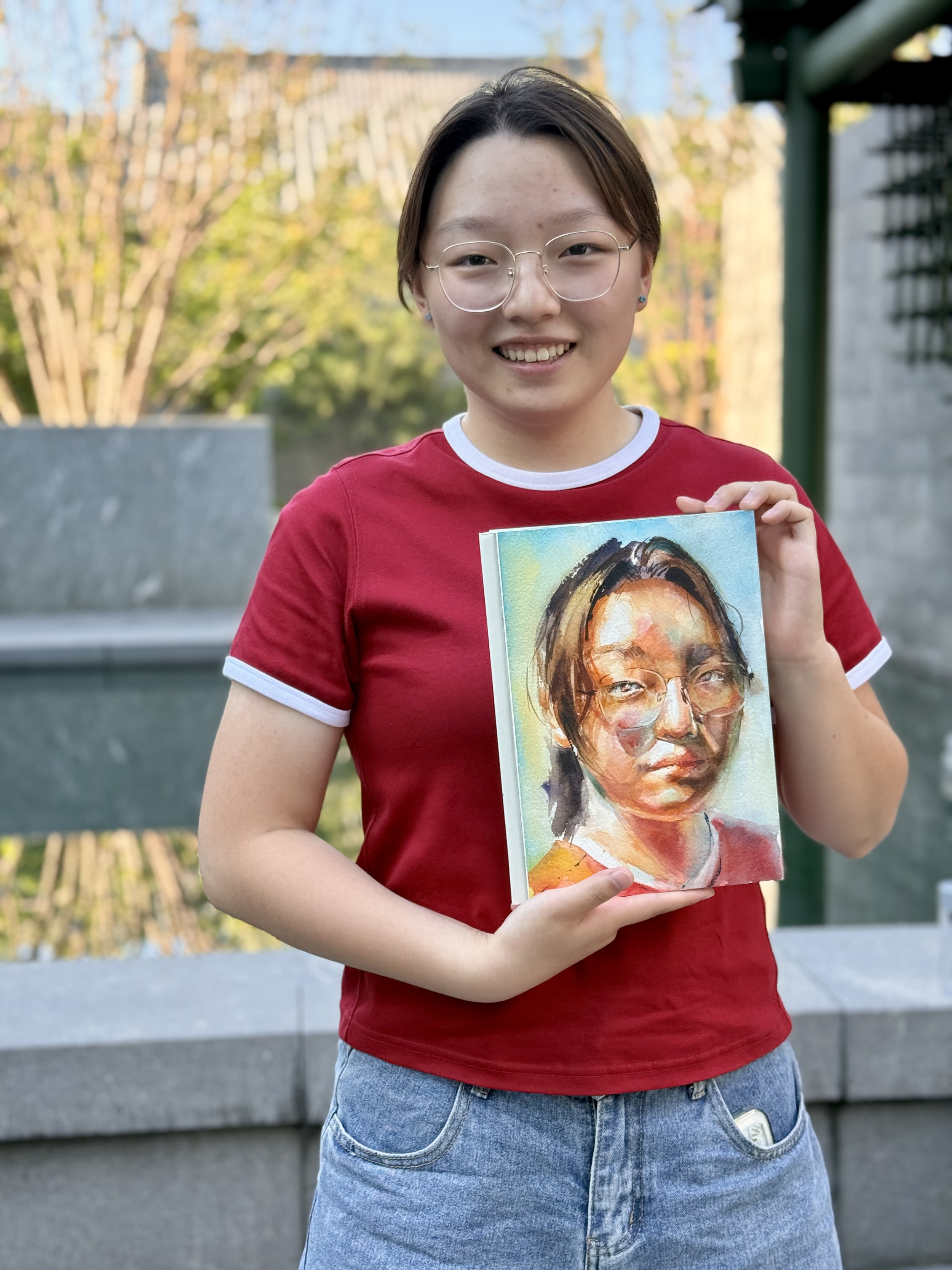 Person wearing glasses and a red shirt holding a colorful watercolor portrait of a woman with glasses.