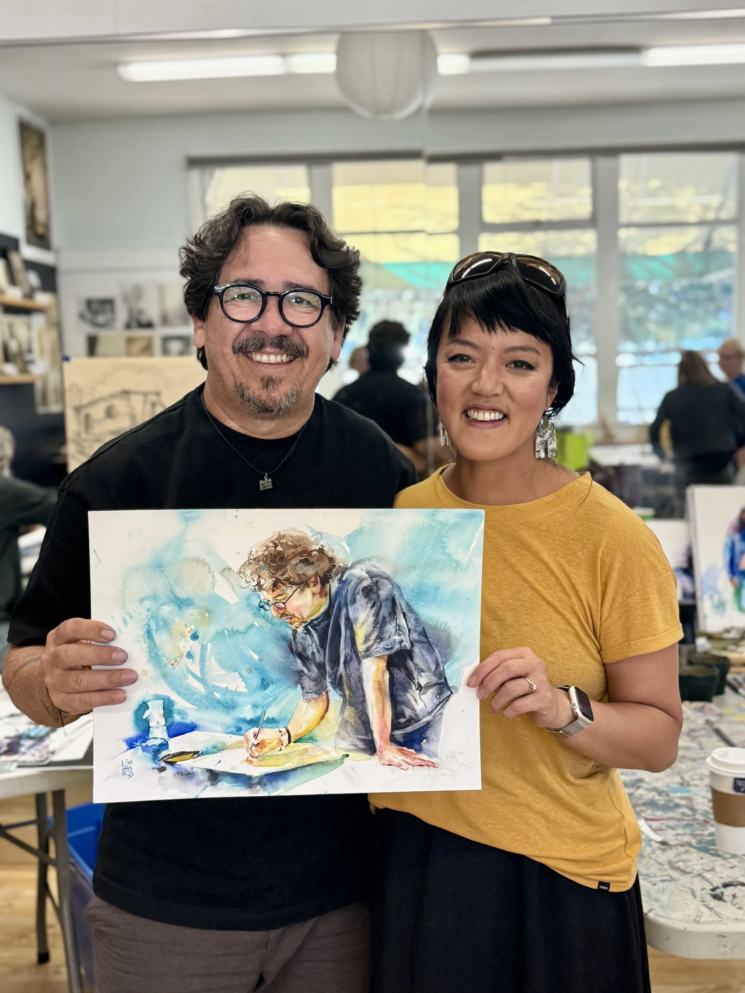 Eudes Correia and Lorraine Simonds holding a watercolor painting together in a well-lit art studio with other art supplies and artworks in the background.