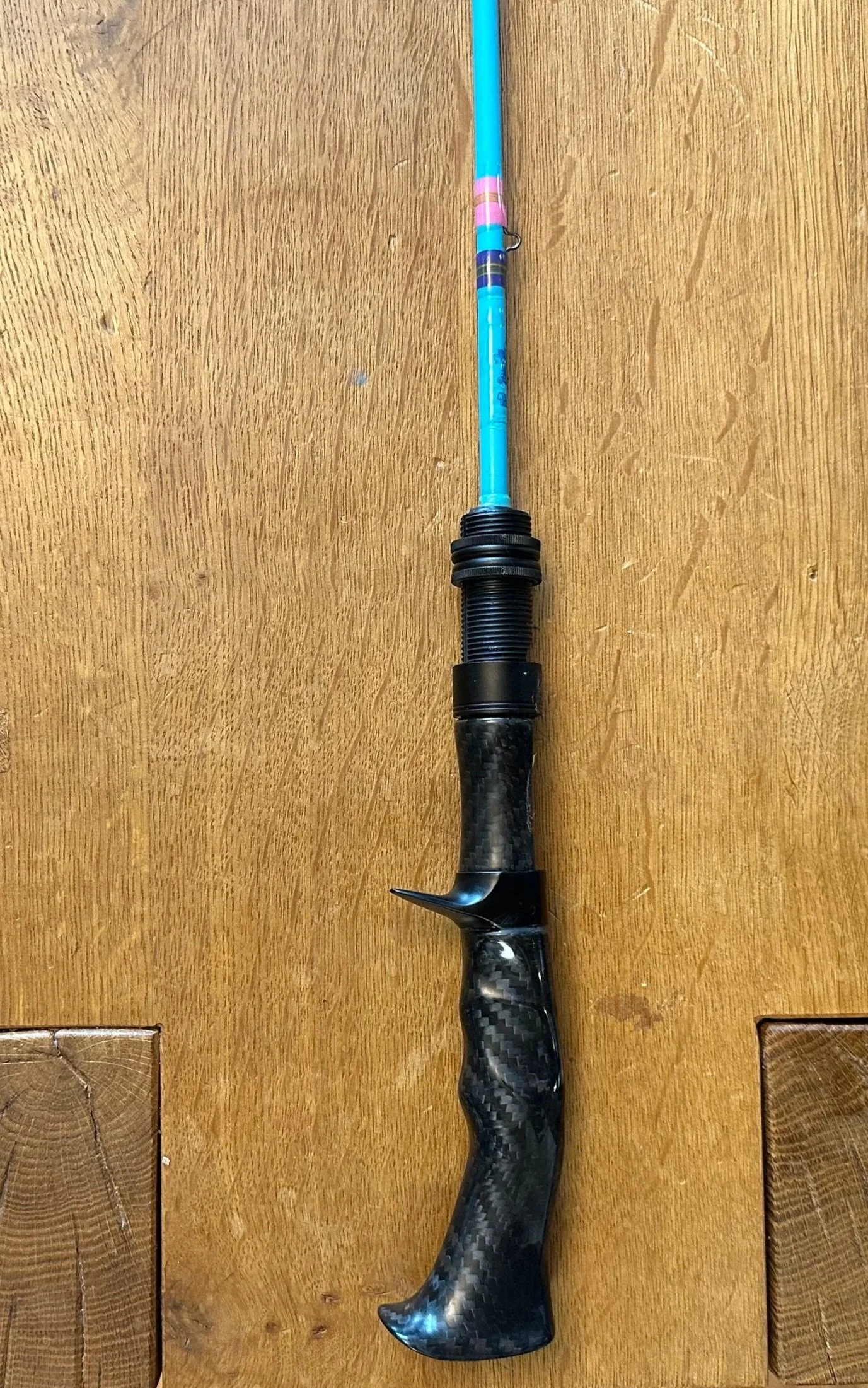 A fishing rod with a black carbon fiber pistol grip handle and blue and pink accents on the upper part, laid out on a wooden surface.
