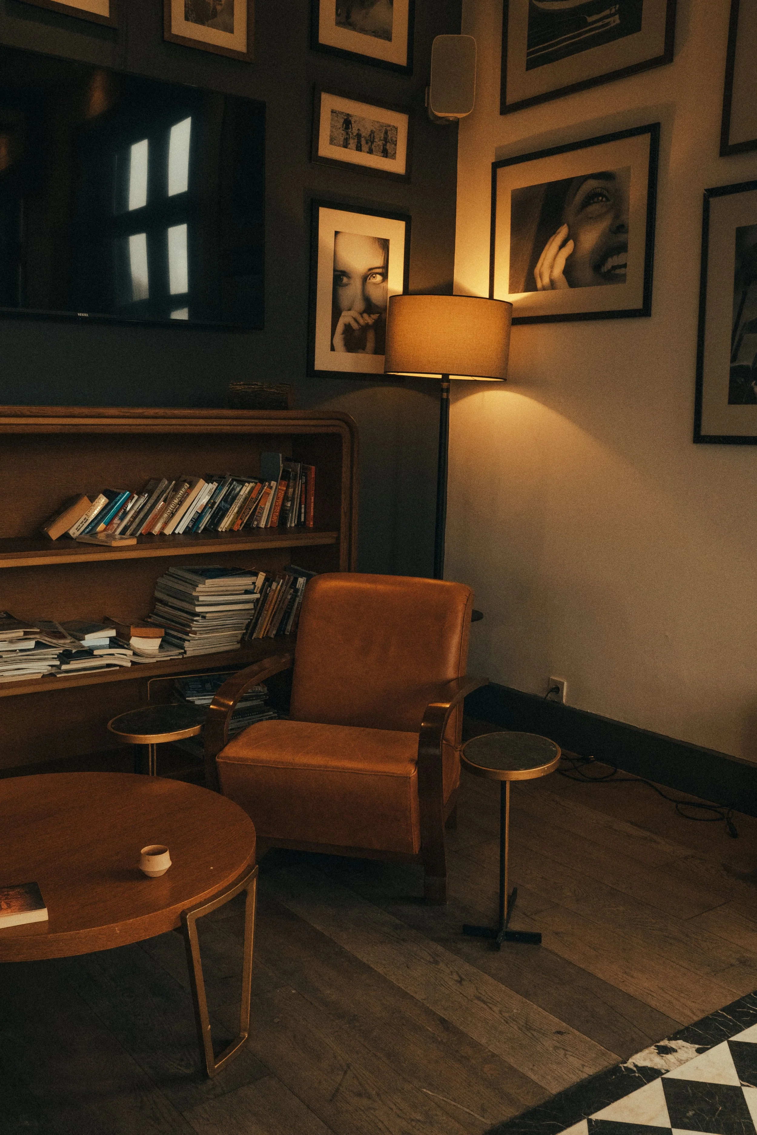 A cozy living room corner with a wooden bookshelf filled with books, a leather armchair, a small round side table, a floor lamp casting warm light, framed black-and-white photographs on the wall, a TV on the wall, and a checkered black and white marble floor border.